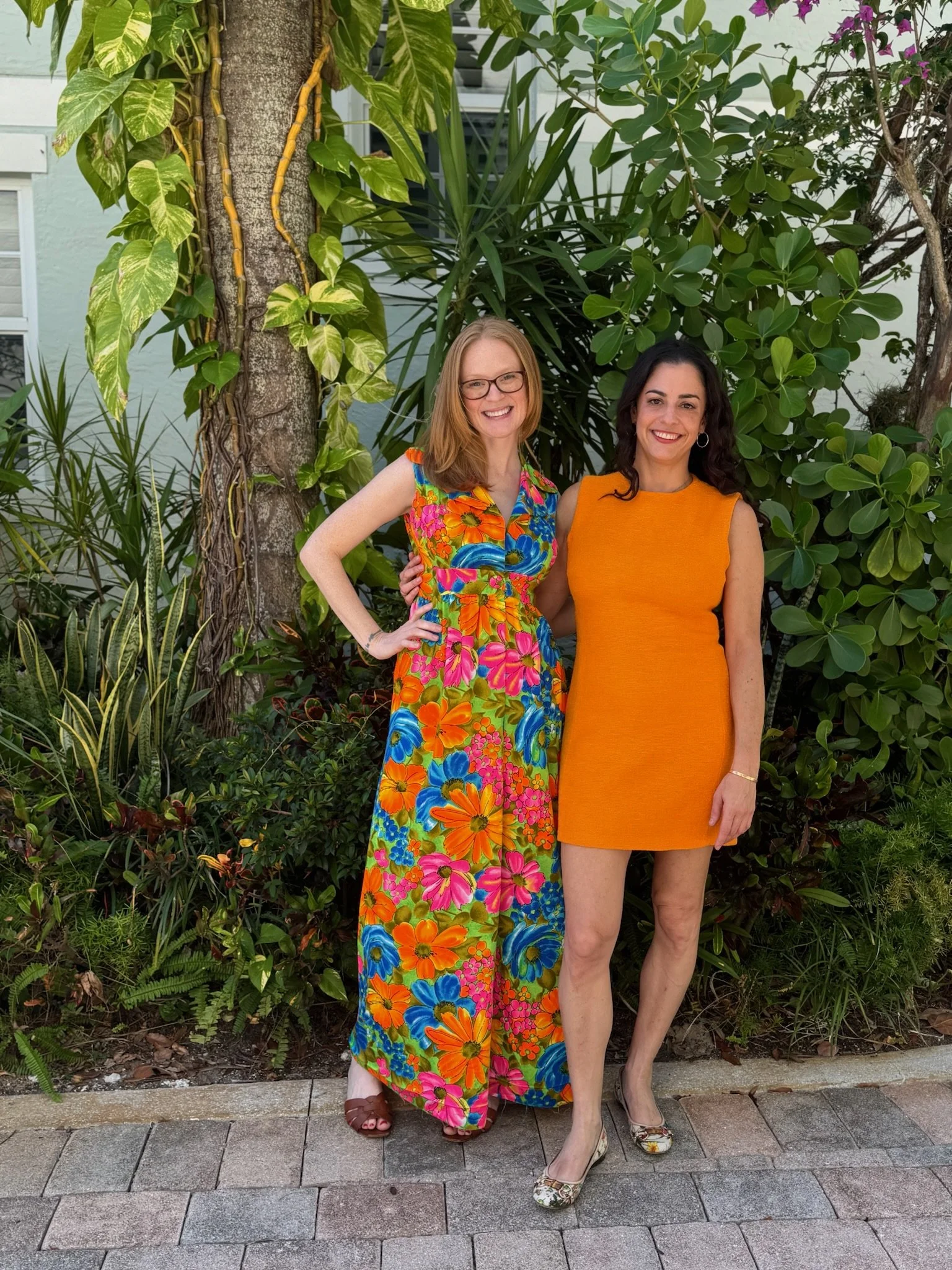 Two ADHD coaches and entrepreneur women standing outdoors in front of lush green plants and trees, smiling at the camera. Build your business with support, understanding, and strategies tailored for your neurodivergent brain.