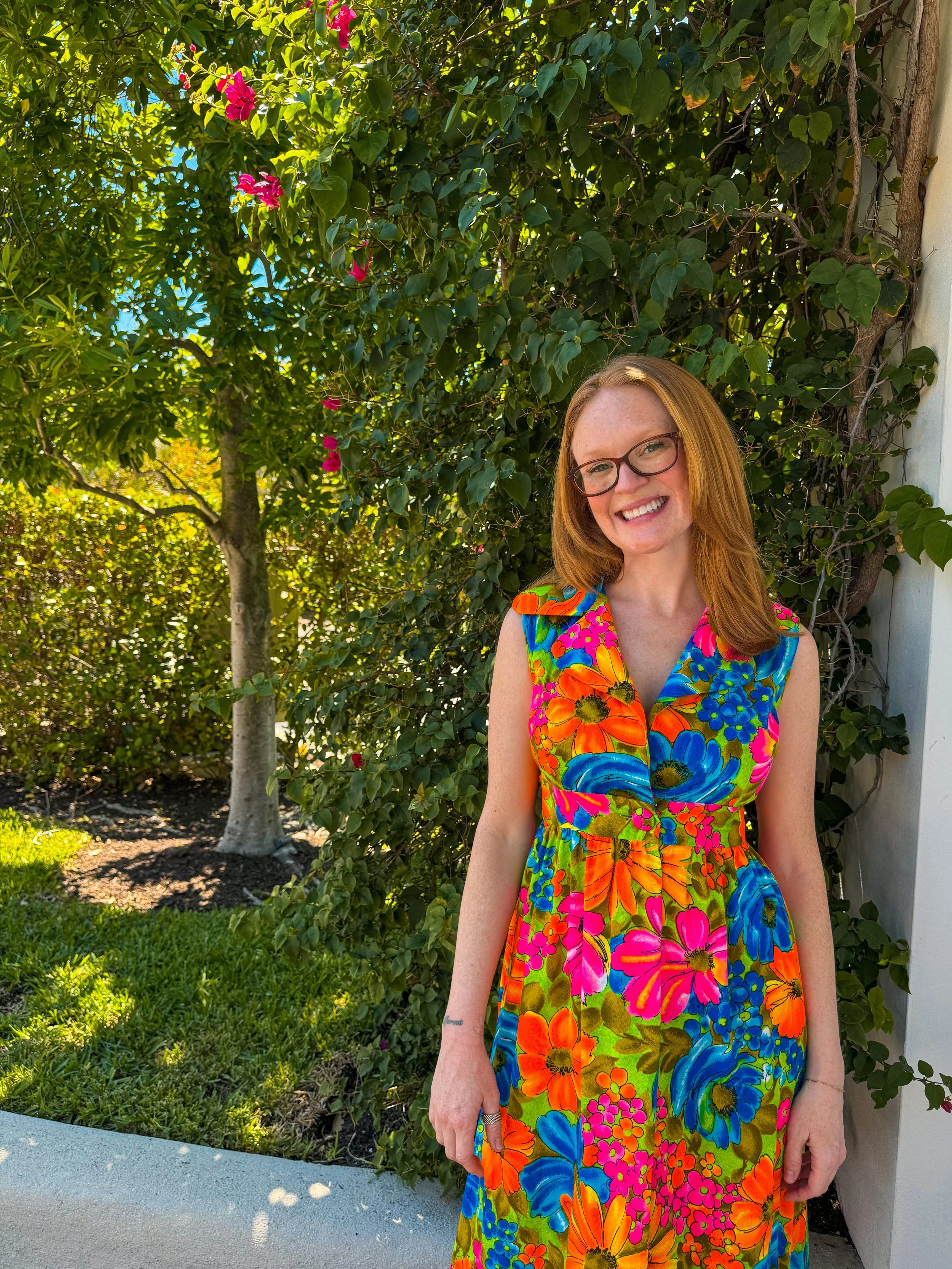 A woman with red hair and glasses smiling outdoors near a green leafy bush with pink flowers, wearing a colorful floral dress.