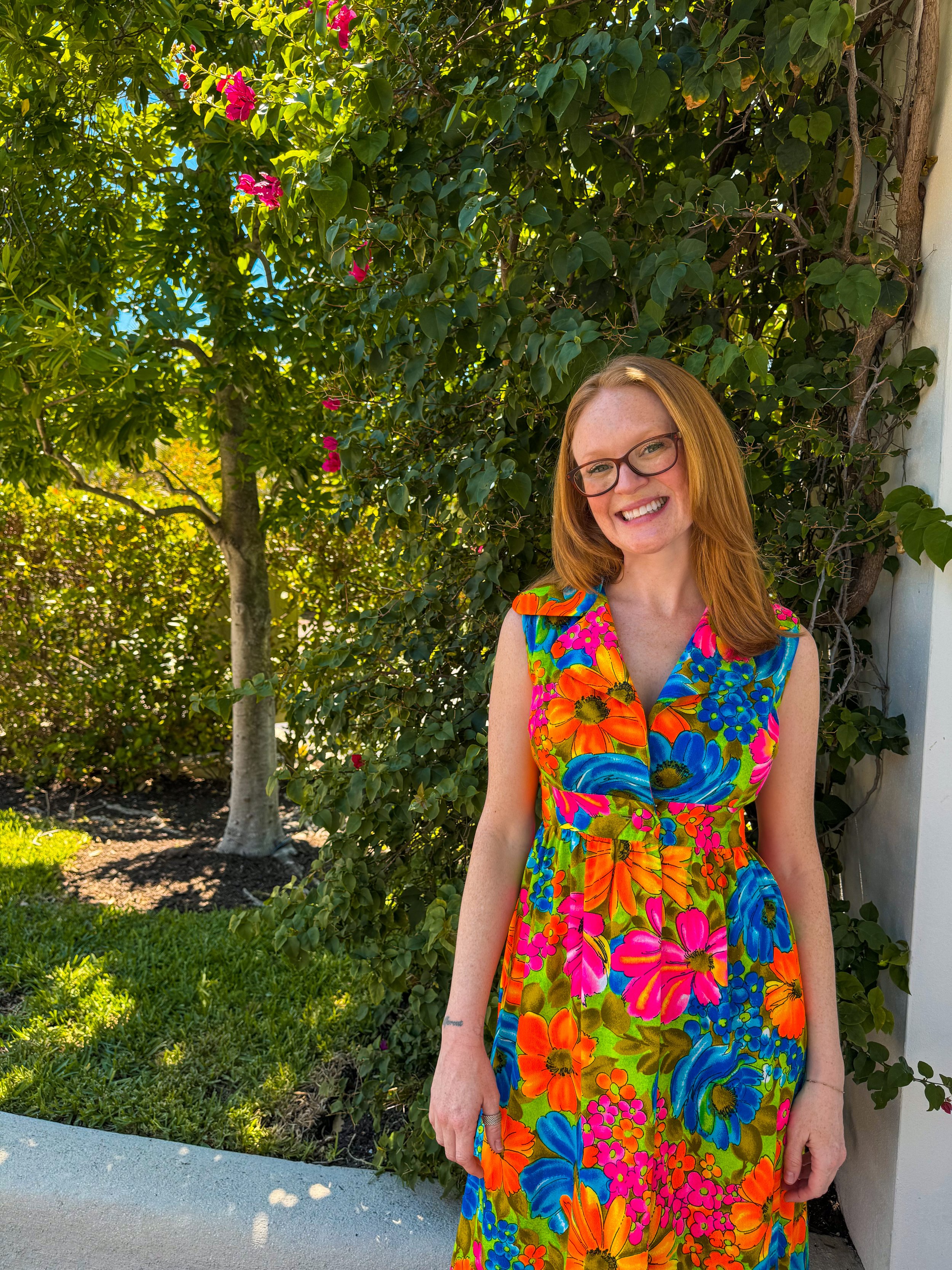 Kate, Neaurospice Girls CEOs, woman with red hair and glasses smiling outdoors near a green leafy bush with pink flowers, wearing a colorful floral dress.
