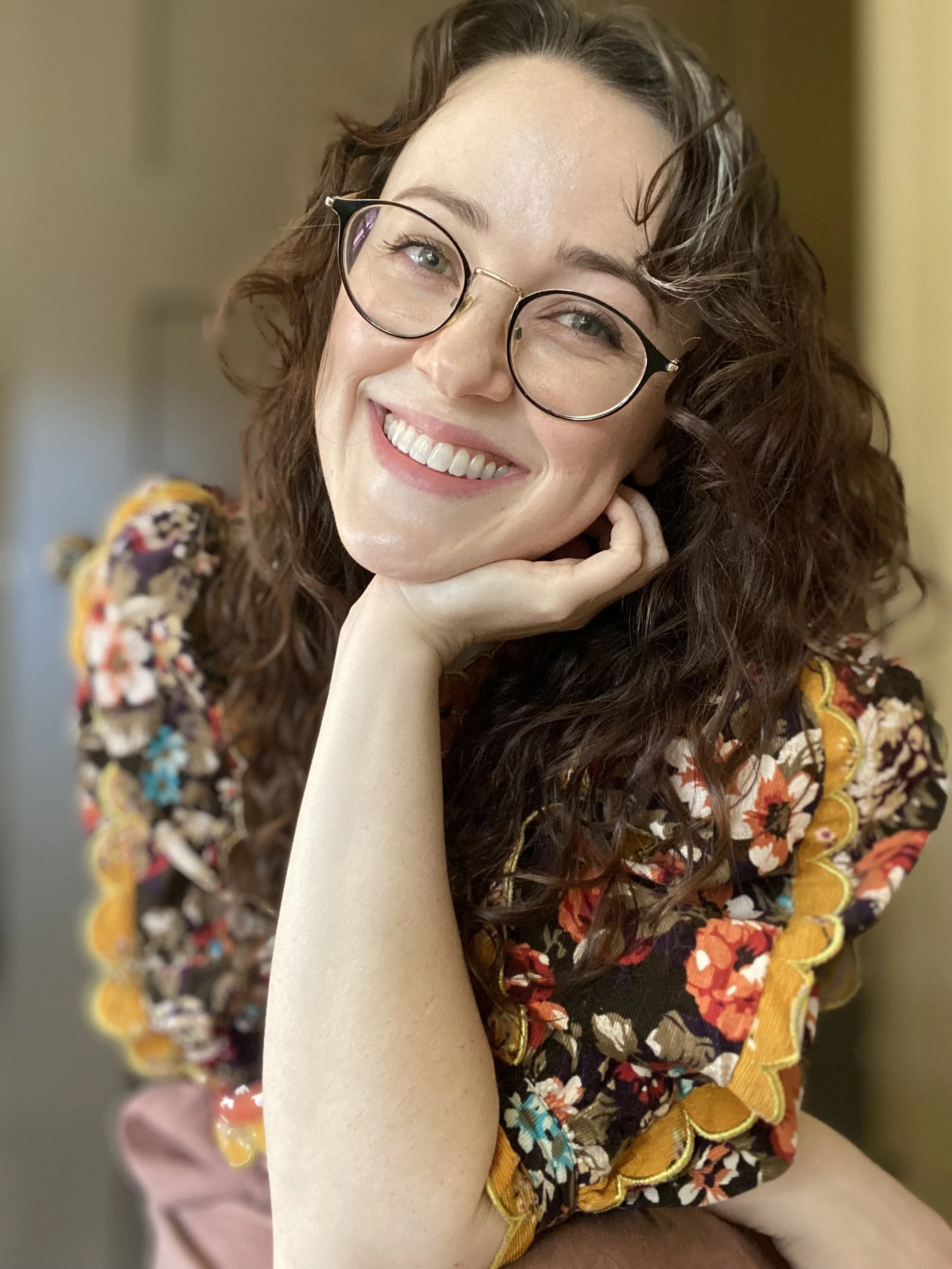 A woman with curly brown hair wearing glasses, smiling, resting her chin on her hand, and dressed in a colorful floral top.