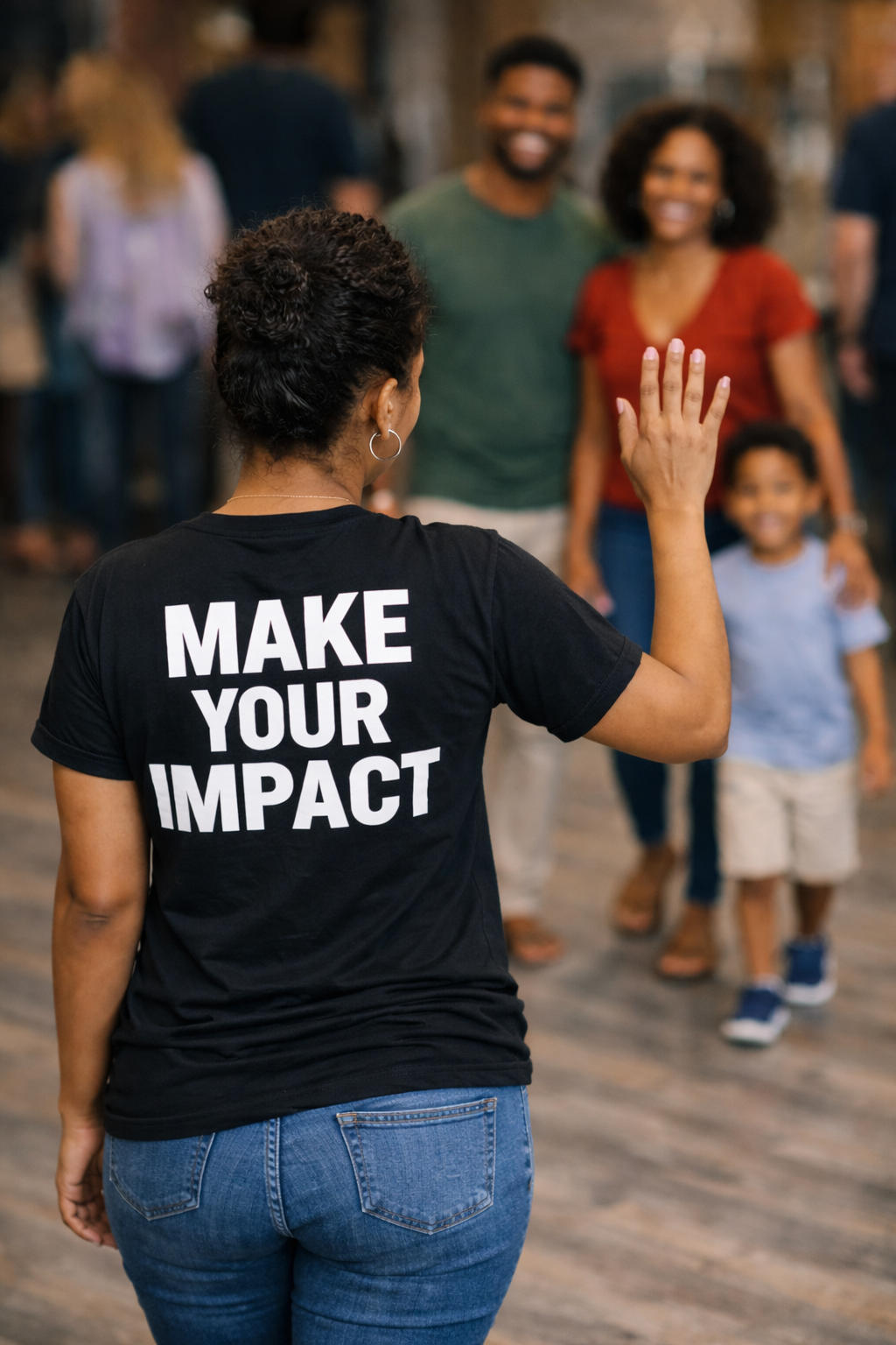 Woman with a shirt that says 'Make Your Impact' waving at a group of smiling people, including adults and a child, in an indoor setting.