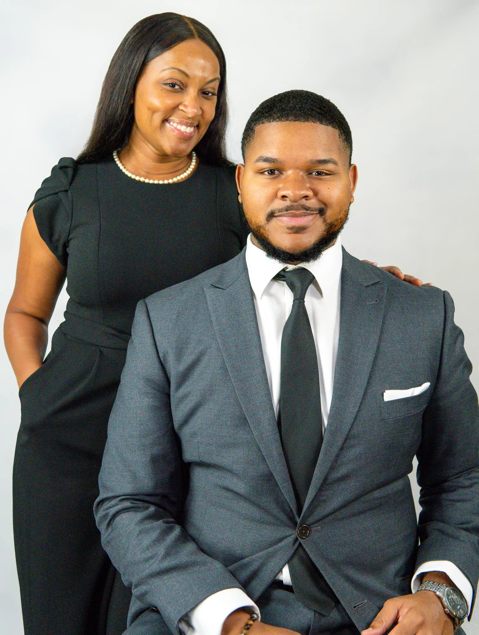A woman and a man dressed in professional attire posing for a photo. The woman is standing behind and slightly to the side of the seated man. She is wearing a black dress and a pearl necklace, smiling warmly. The man is wearing a gray suit, white shirt, and black tie, looking confident with a slight smile.