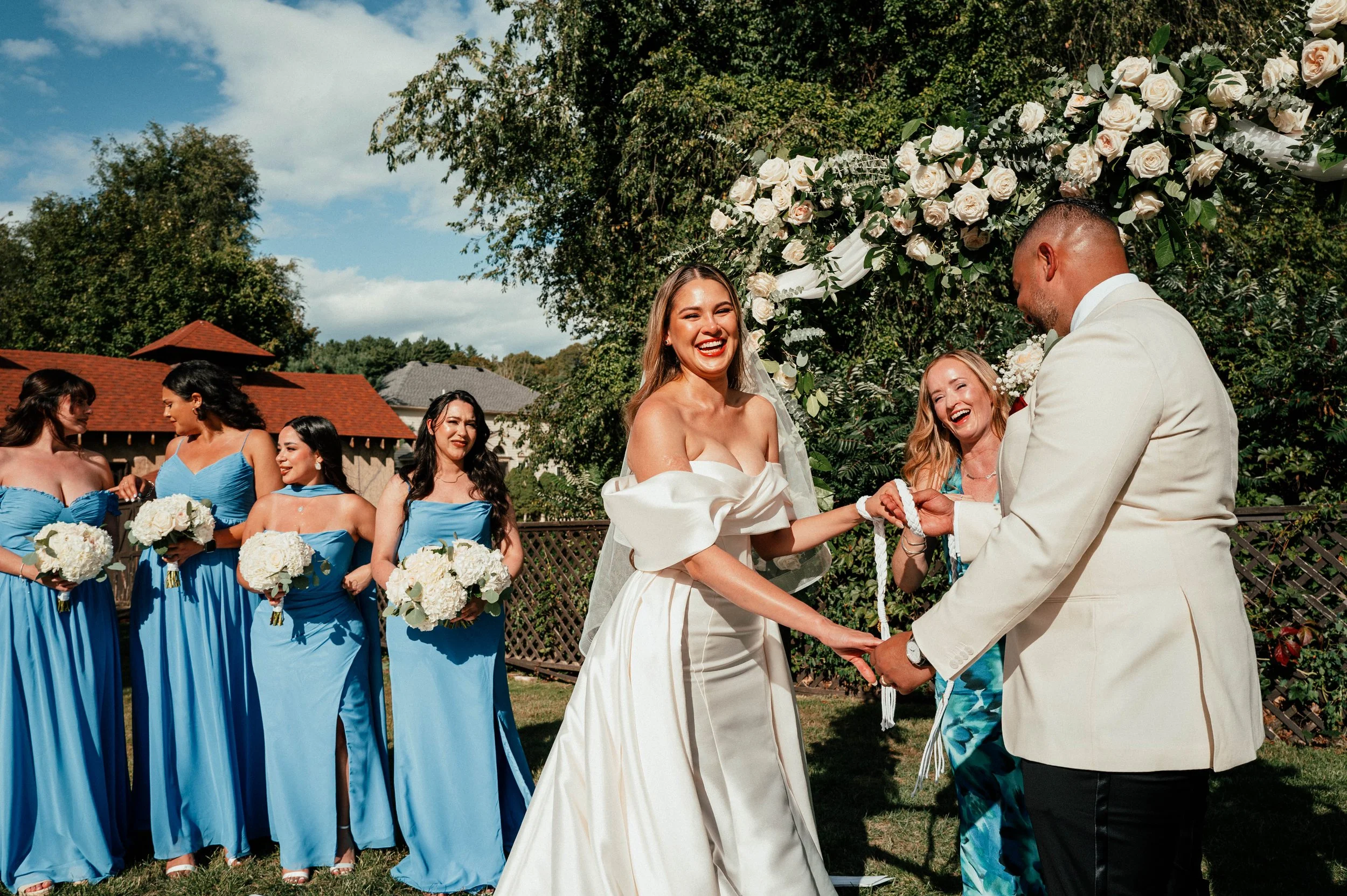 Bride and groom exchanging vows outdoors with bridesmaids in blue dresses holding bouquets in the background, greenery, and a floral arch.