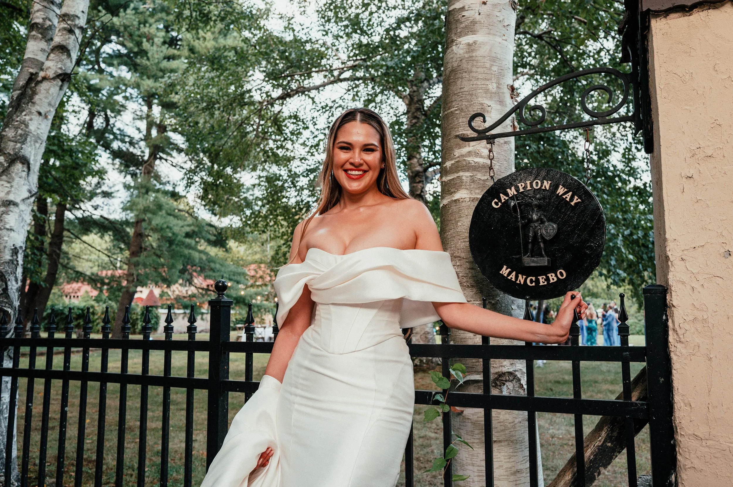 A smiling woman in a strapless white dress stands outdoors near a tree, holding a black sign that says 'Champion Way Mancebo' with a graphic of a figure holding a trophy.
