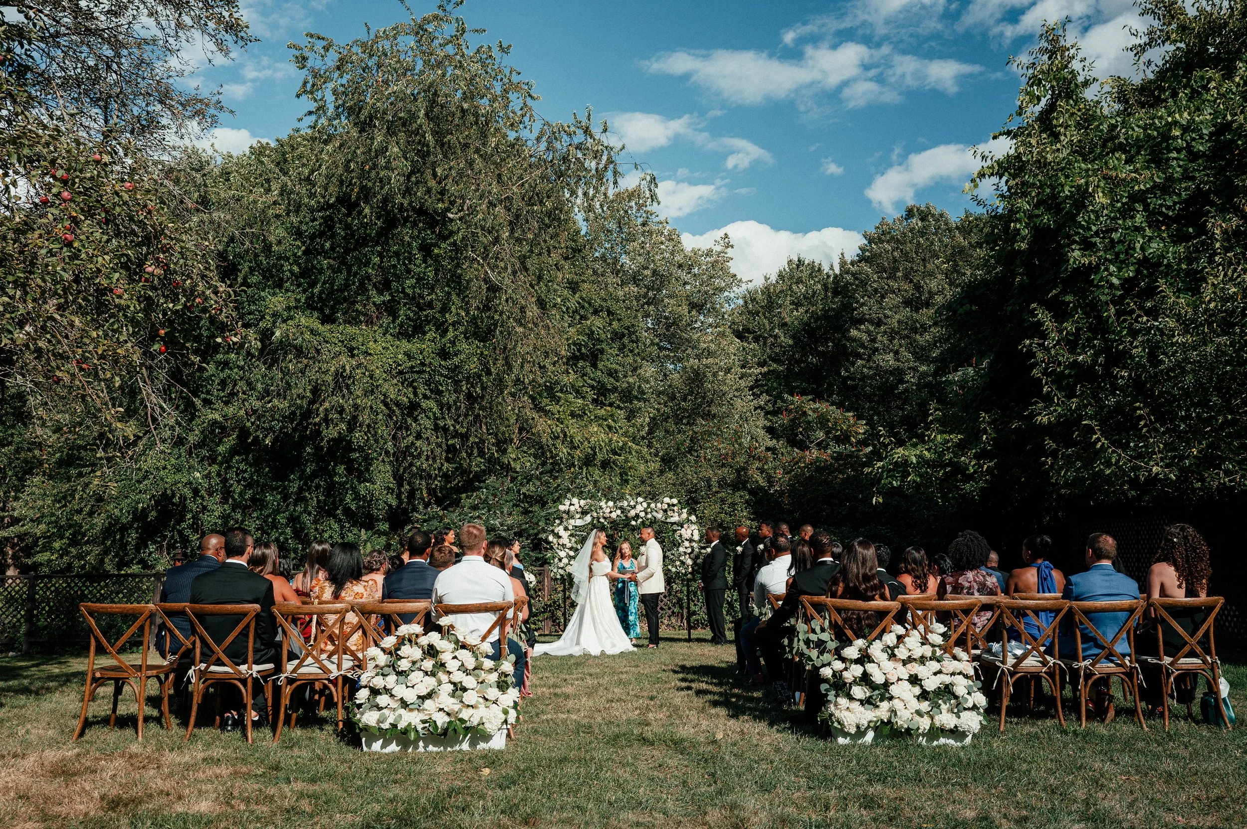 Outdoor wedding ceremony with a bride and groom facing each other under a floral arch, surrounded by seated guests, amidst greenery and a bright blue sky.