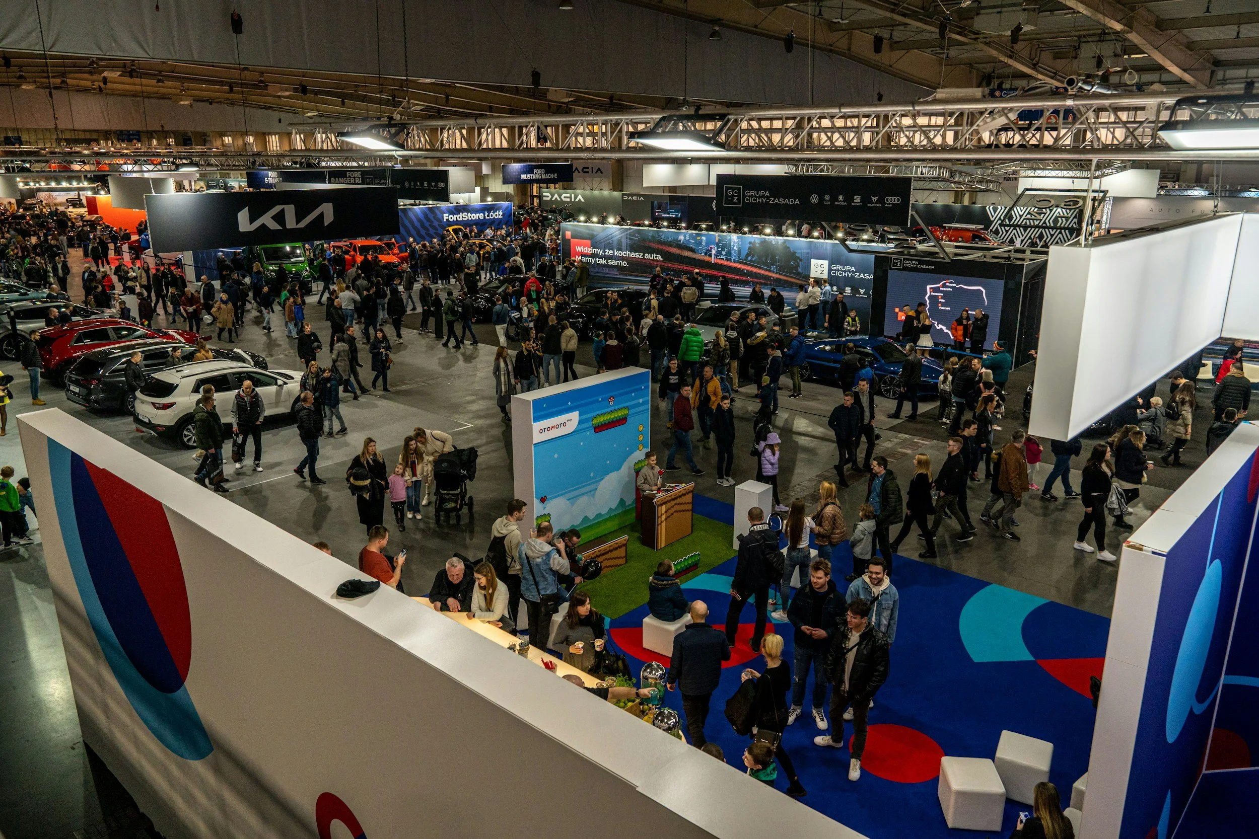 Interior of an auto show with a large crowd of people, displaying various cars, booths, and branded signs.