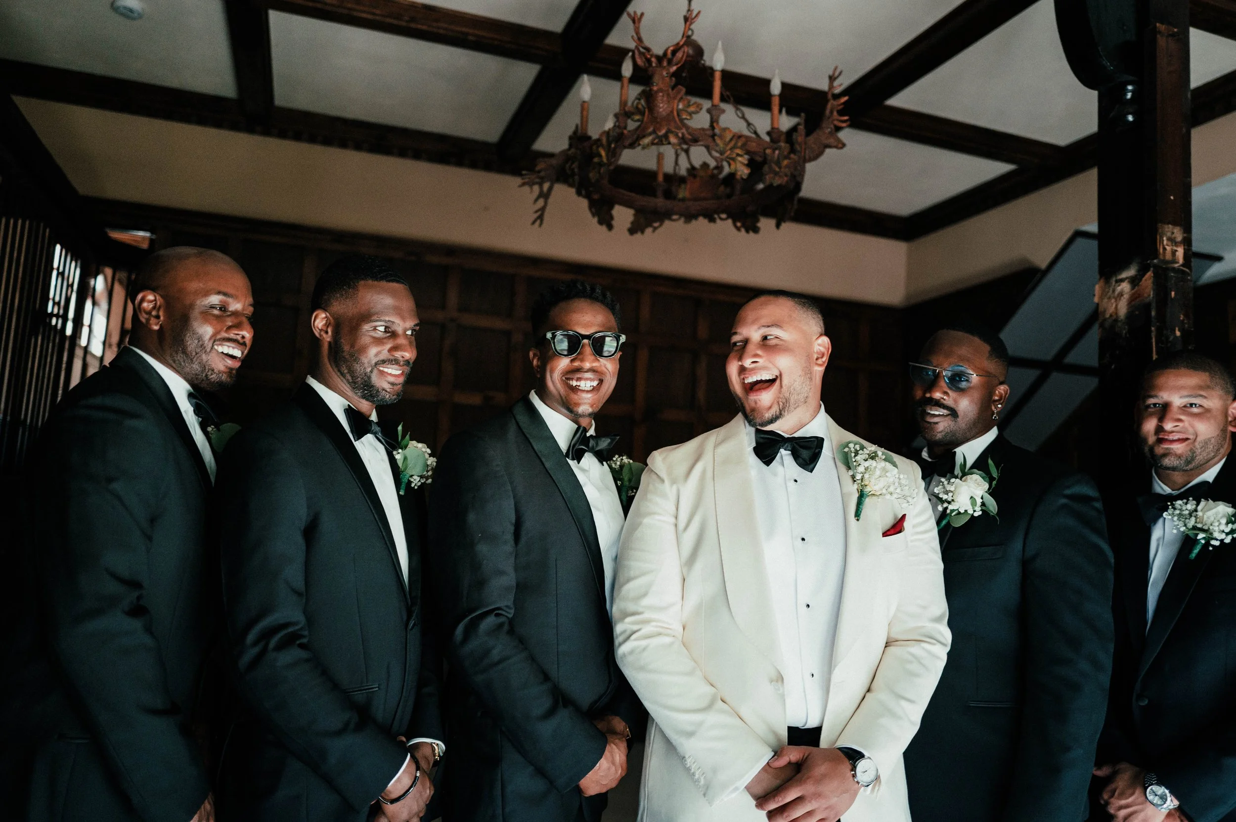 A group of seven men in tuxedos, with the groom in a cream tuxedo, smiling and looking at each other indoors under a chandelier.