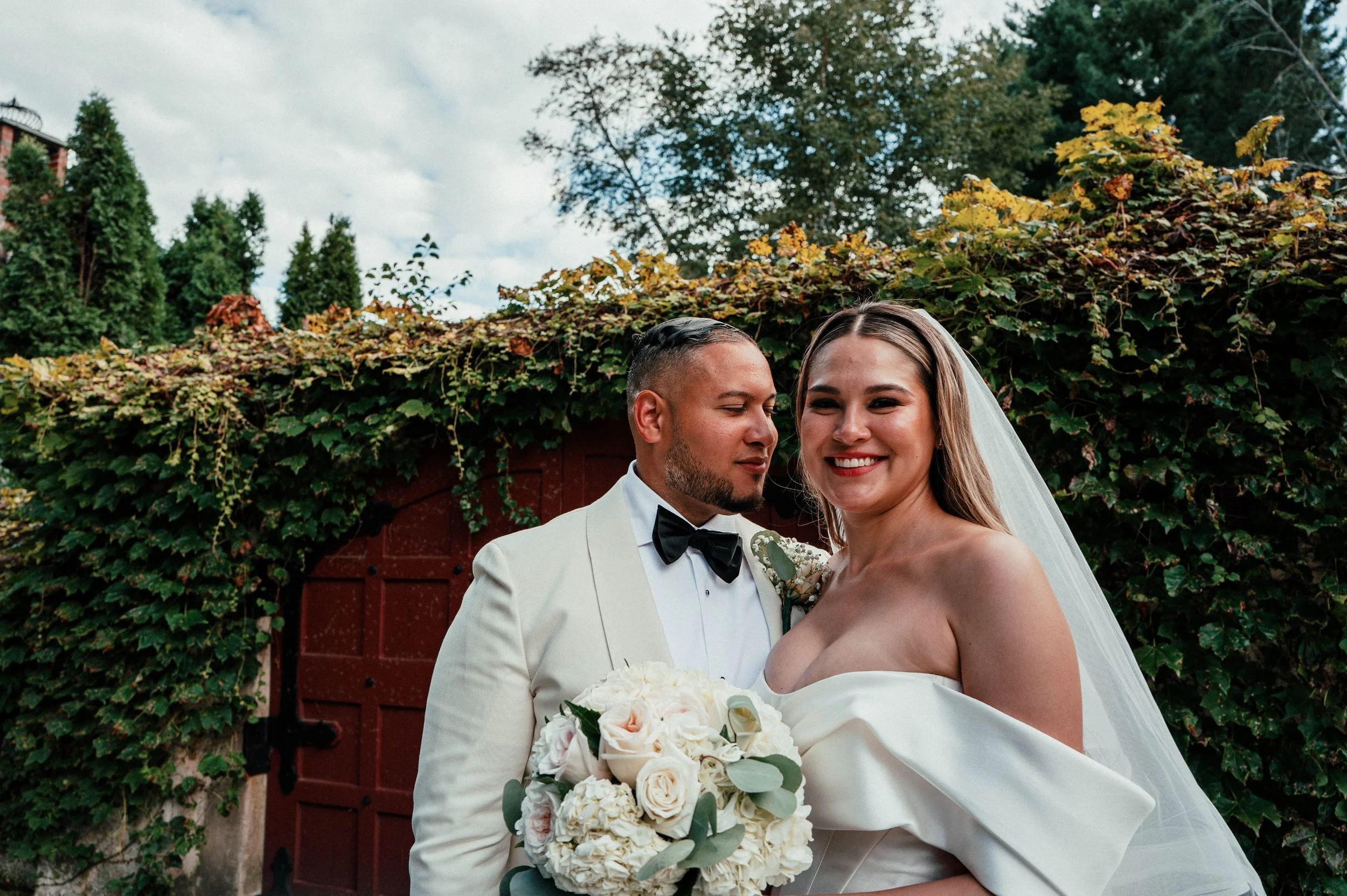 A newlywed couple on their wedding day, standing outdoors in front of a red door and green foliage. The groom, dressed in a white tuxedo with a black bow tie, stands close to the bride. The bride, wearing a strapless wedding gown and veil, holds a bo