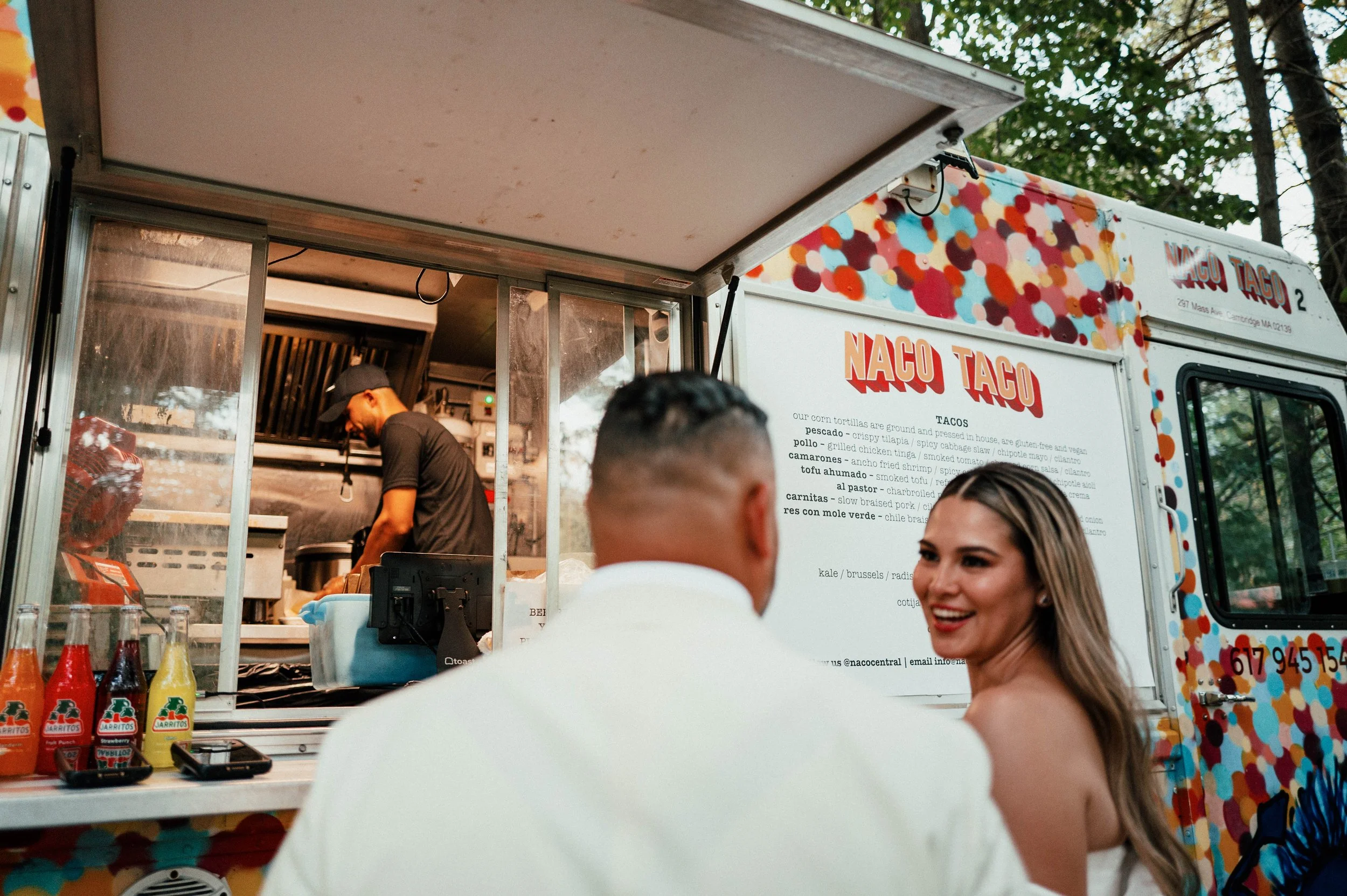 A food truck named NACO TACO serving tacos with a menu on the side. Two customers, a man in a white shirt and a woman with long hair, are talking in front of the truck. The woman is smiling.