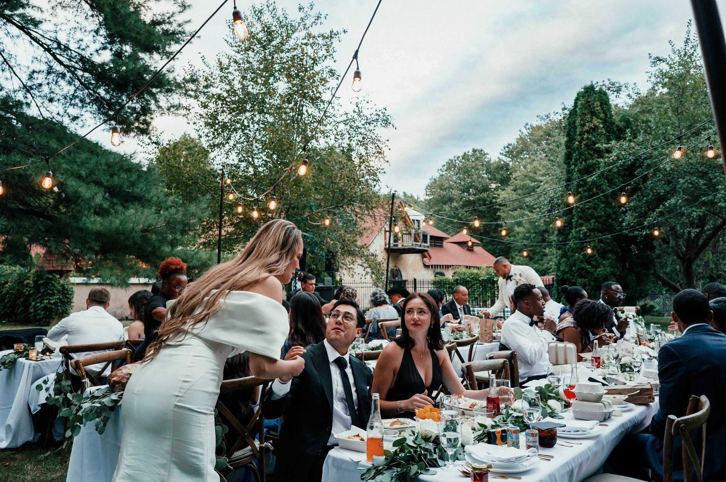 People dressed in formal attire enjoying a wedding reception outdoors. A woman in a white dress is leaning over to speak with a man in a dark suit and glasses, both seated at a long table decorated with greenery and drinks. Overhead string lights ill