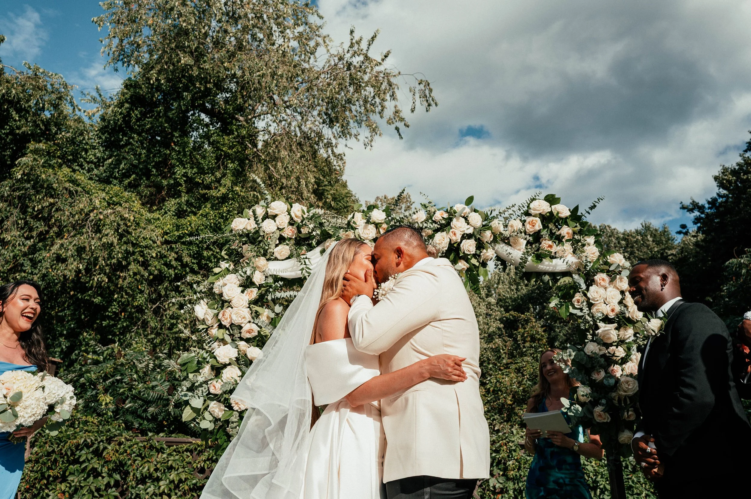 A wedding ceremony outdoors with a couple kissing under a floral arch, surrounded by friends and greenery.
