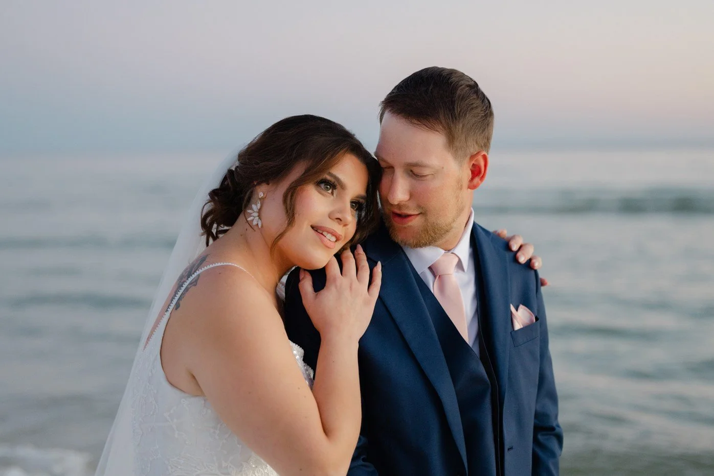 A bride and groom standing close together on a beach during sunset, with the bride resting her head on the groom's shoulder and smiling softly.
