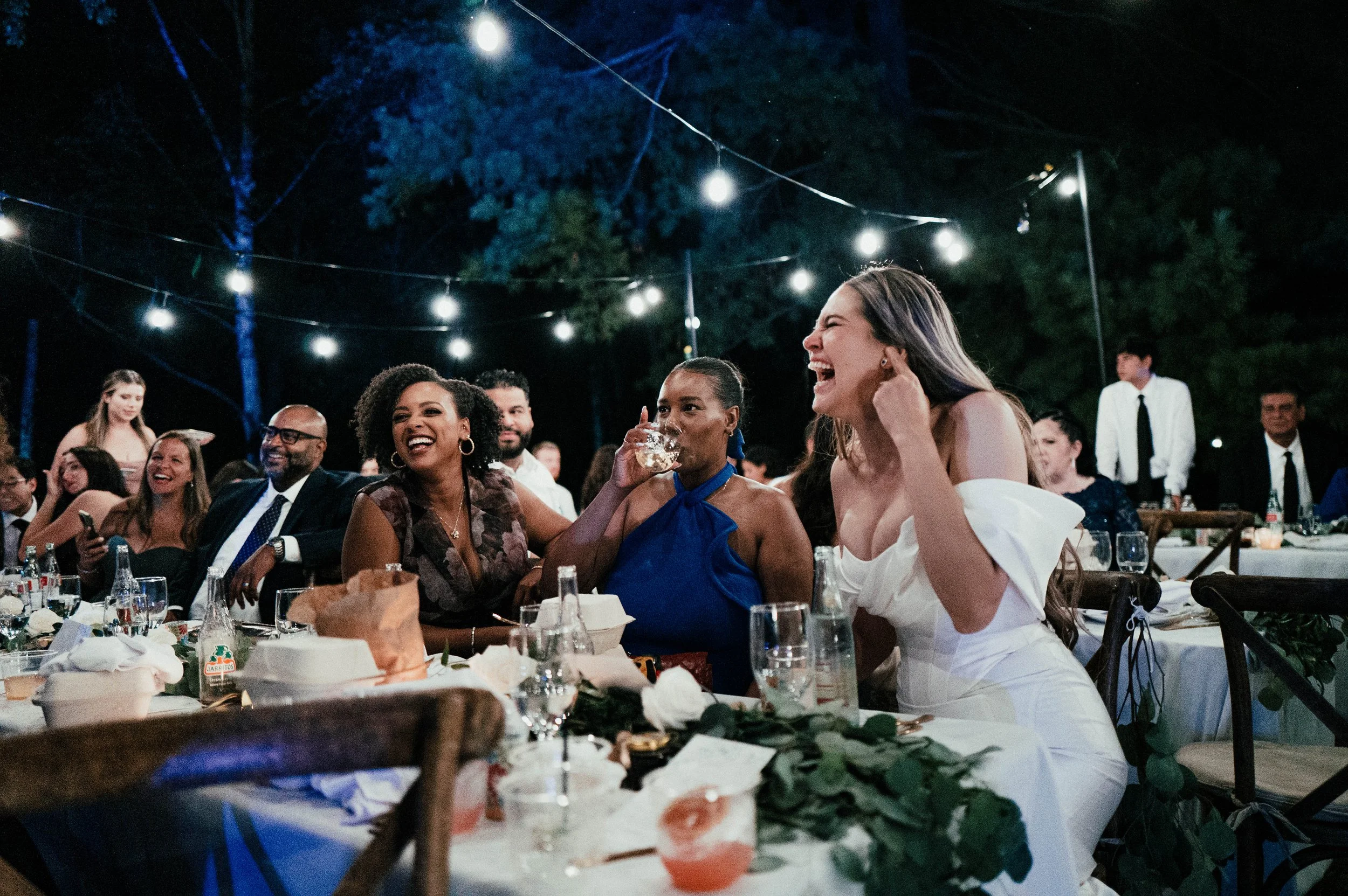 People enjoying a nighttime outdoor celebration with string lights overhead, seated at decorated tables, laughing and socializing.