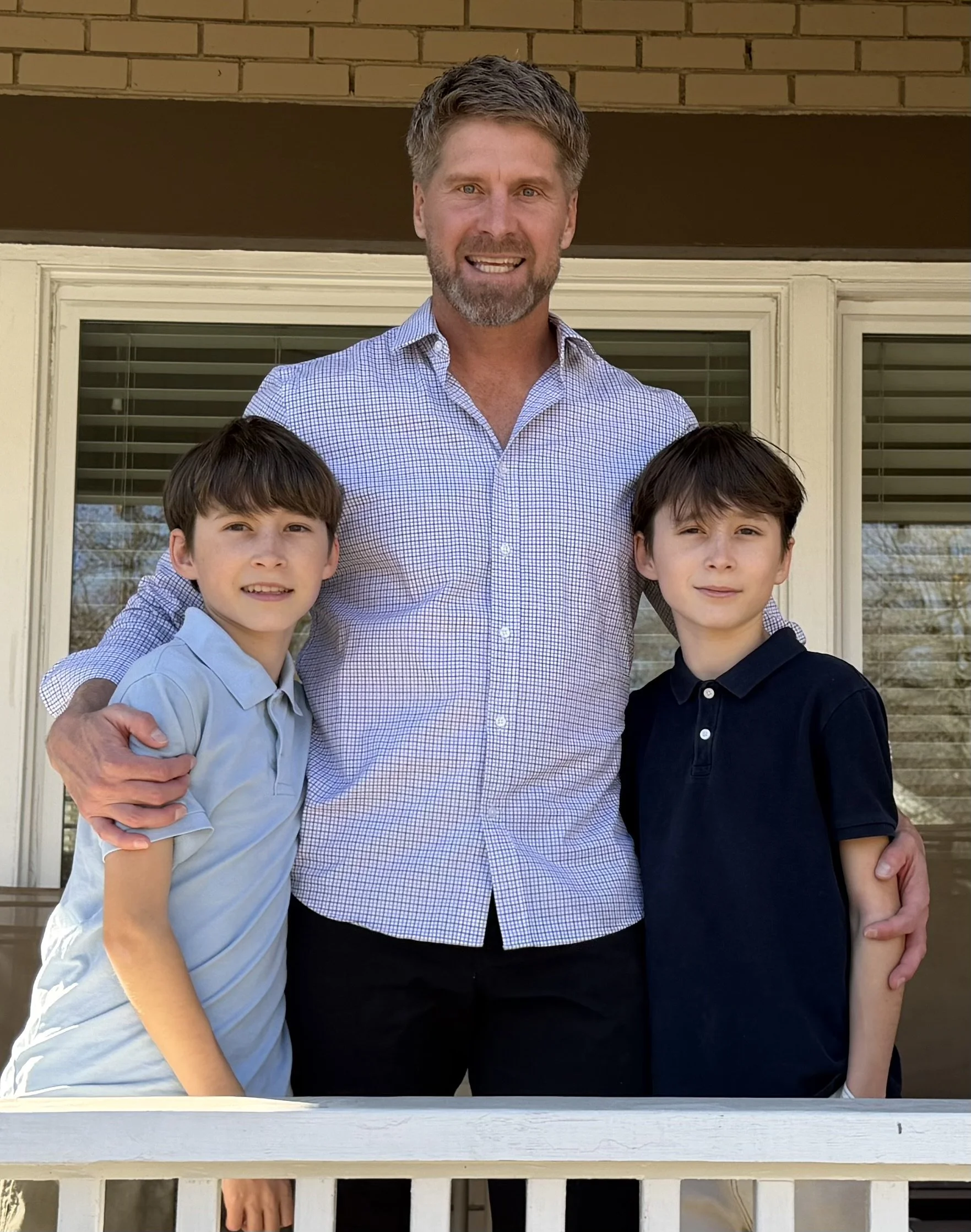A man with two boys standing outside on a balcony in front of a house, smiling at the camera.