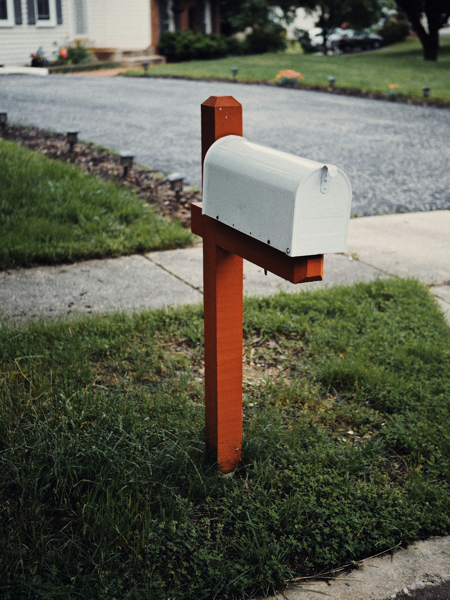White mailbox mounted on a red wooden post in a front yard with green grass, sidewalk, and street in the background.