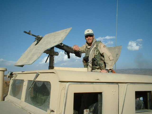 A soldier in military gear standing inside a tan armored vehicle, operating a mounted machine gun, with a clear blue sky in the background.