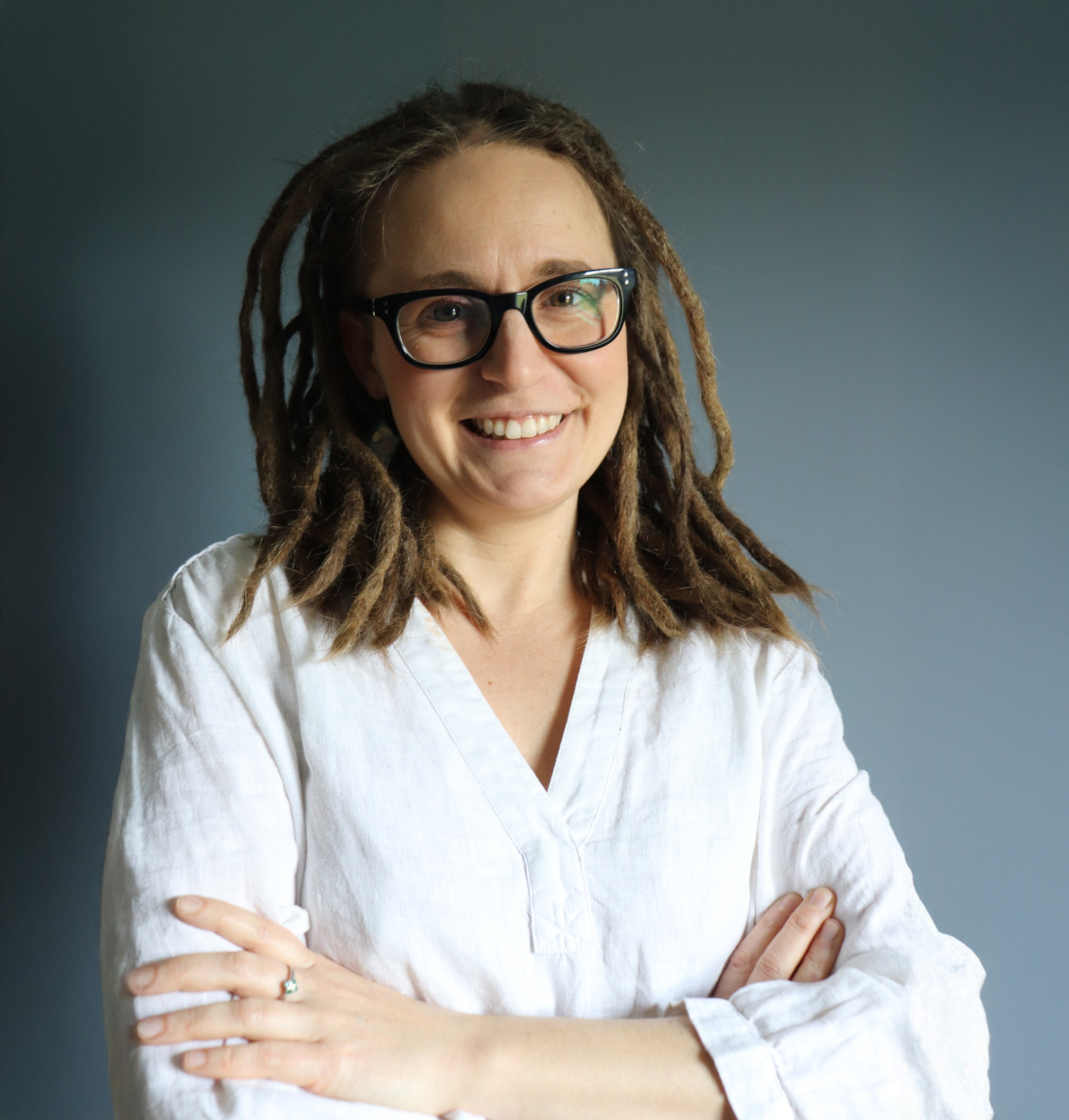 A woman with dreadlocks, glasses, and a white shirt smiling with arms crossed against a blue background.