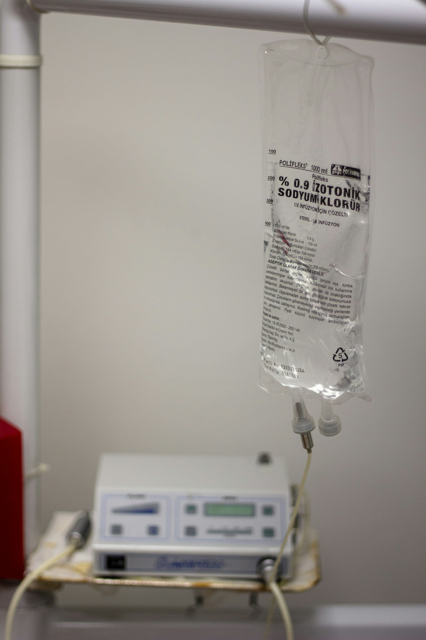 An intravenous (IV) fluid bag labeled in a foreign language hanging connected to a medical infusion pump, with a white wall in the background.