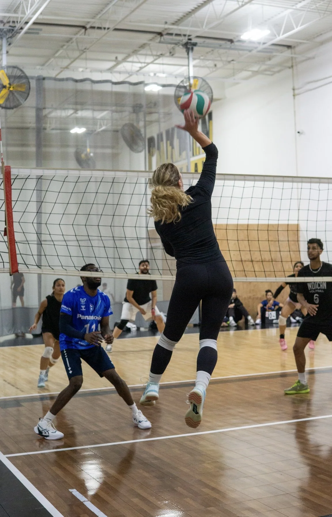 A woman jumps to block a volleyball at an indoor volleyball game, surrounded by players on both teams.