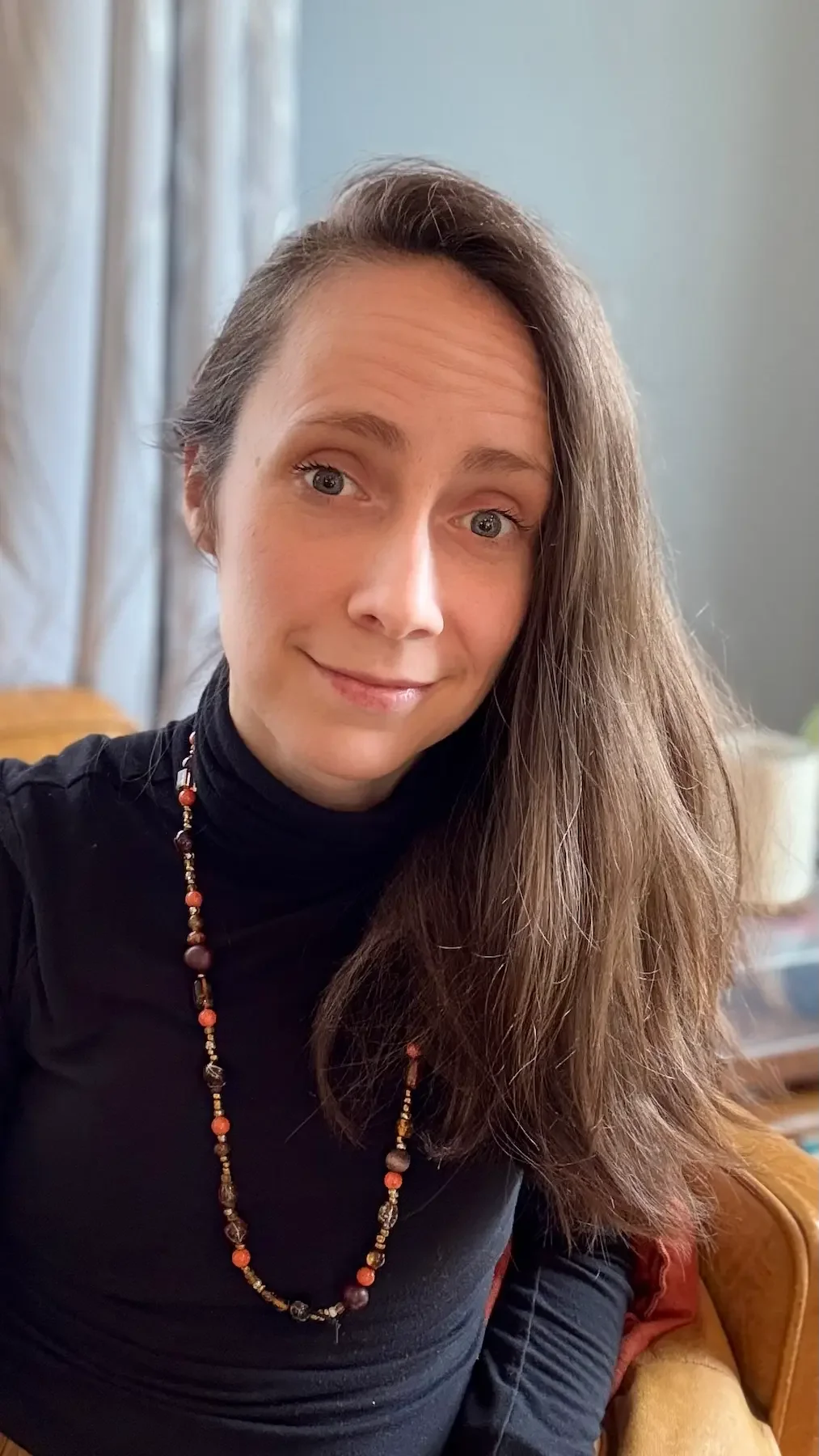 A woman with long brown hair, wearing a black turtleneck and a beaded necklace, smiling while sitting in a room with light-colored curtains and a green plant in the background.