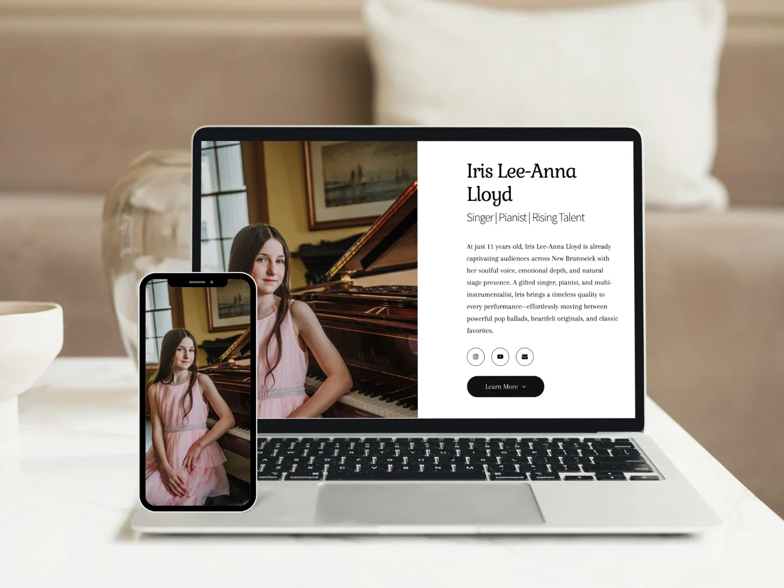 A laptop and a smartphone on a white table displaying a profile of Iris Lee-Anna Lloyd, a young singer and pianist, with her photo next to her biography. The background includes a glass vase, a bowl, and a neutral-colored wall.