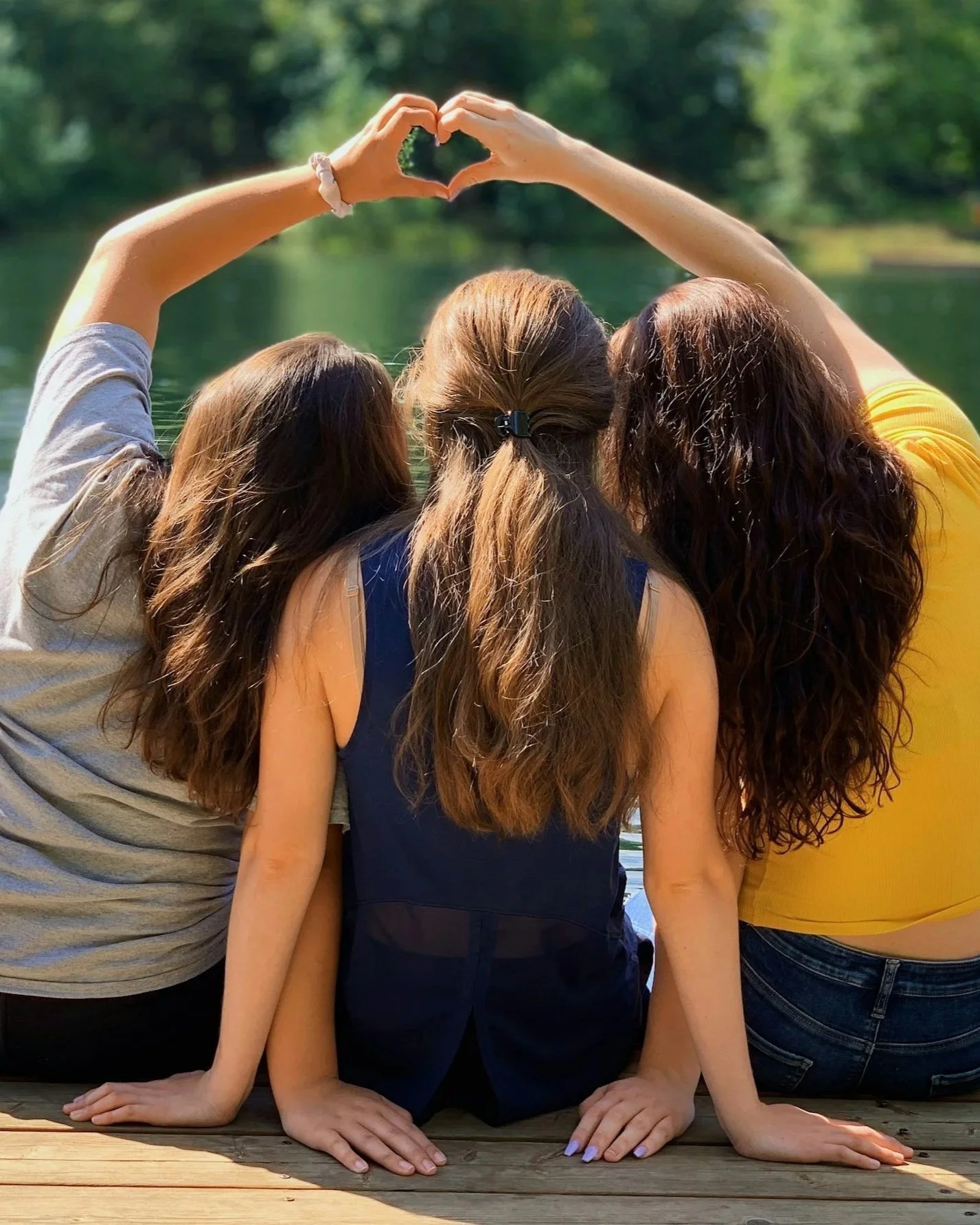 Three girls sitting with their backs to the camera on a wooden dock near a lake, forming a heart shape with their hands above their heads. Friendships at camp are amazing.