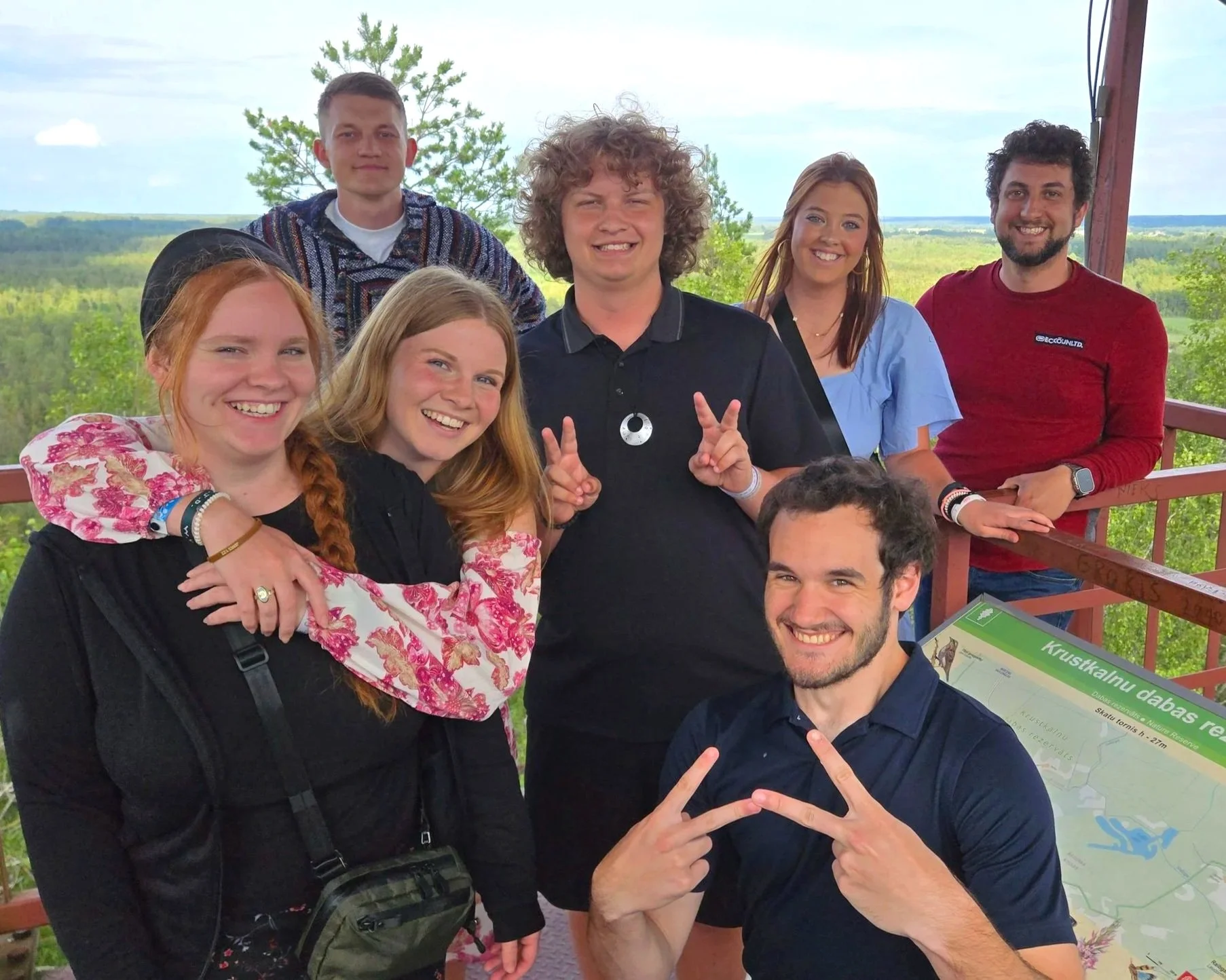 Group of eight people smiling and posing for a photo outdoors on a viewing platform with a lush green landscape in the background. This is what experiencing friendship together while making a difference in the world.