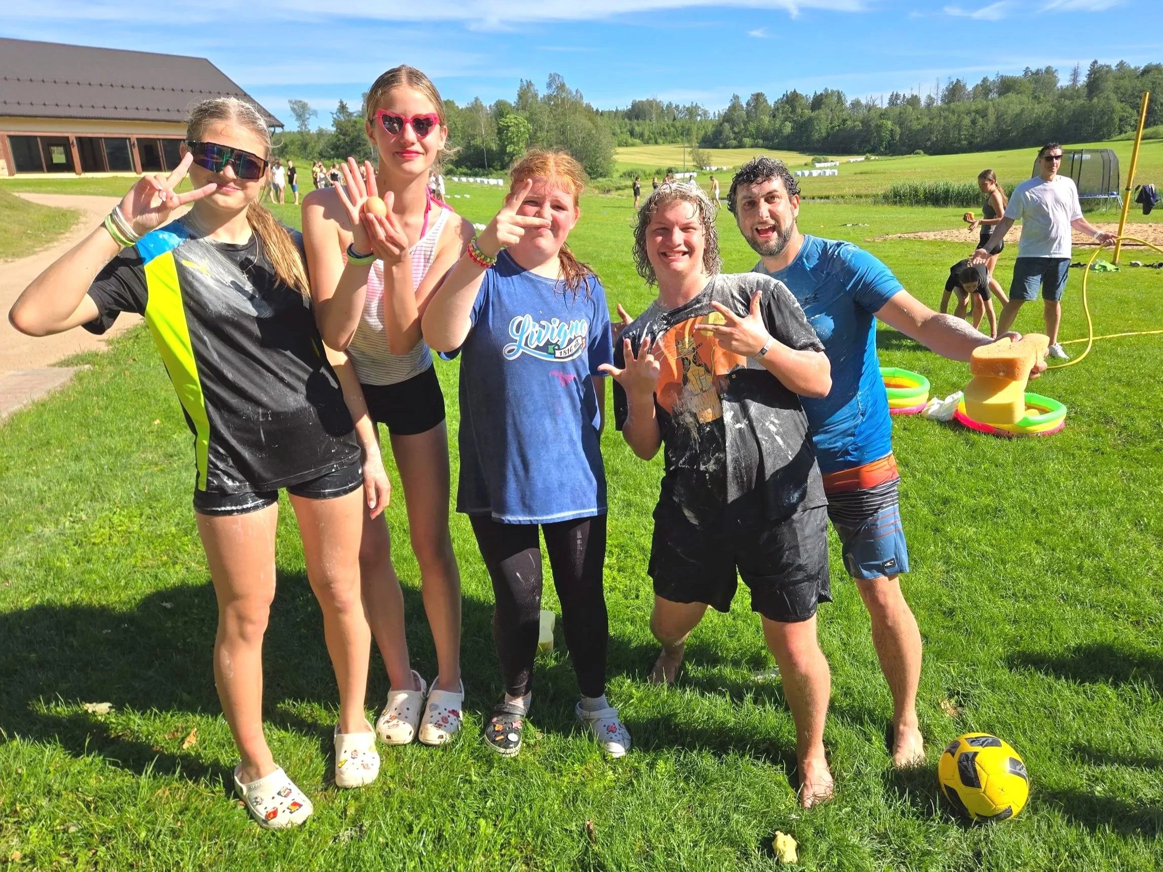 Group of six friends outdoors on grass, throwing water balloons, with a soccer ball nearby, in a sunny park with a building and other people in the background. Camp gets a little crazy!