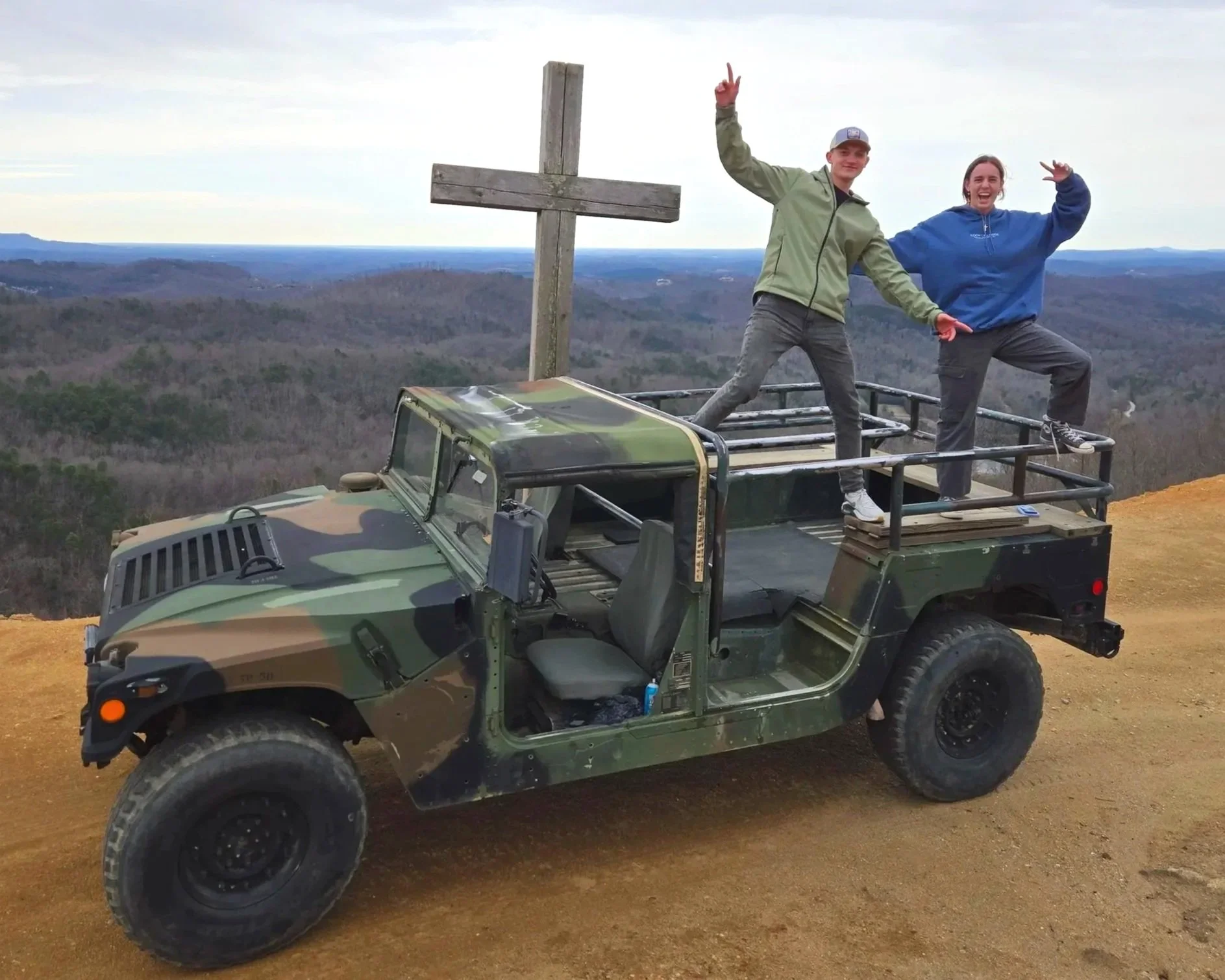 Two young people standing on the back of a camouflaged military-style vehicle near a large wooden cross at a scenic overlook with a vast landscape in the background. When you bring a European to America, nothing beats a hill climb in hummer.