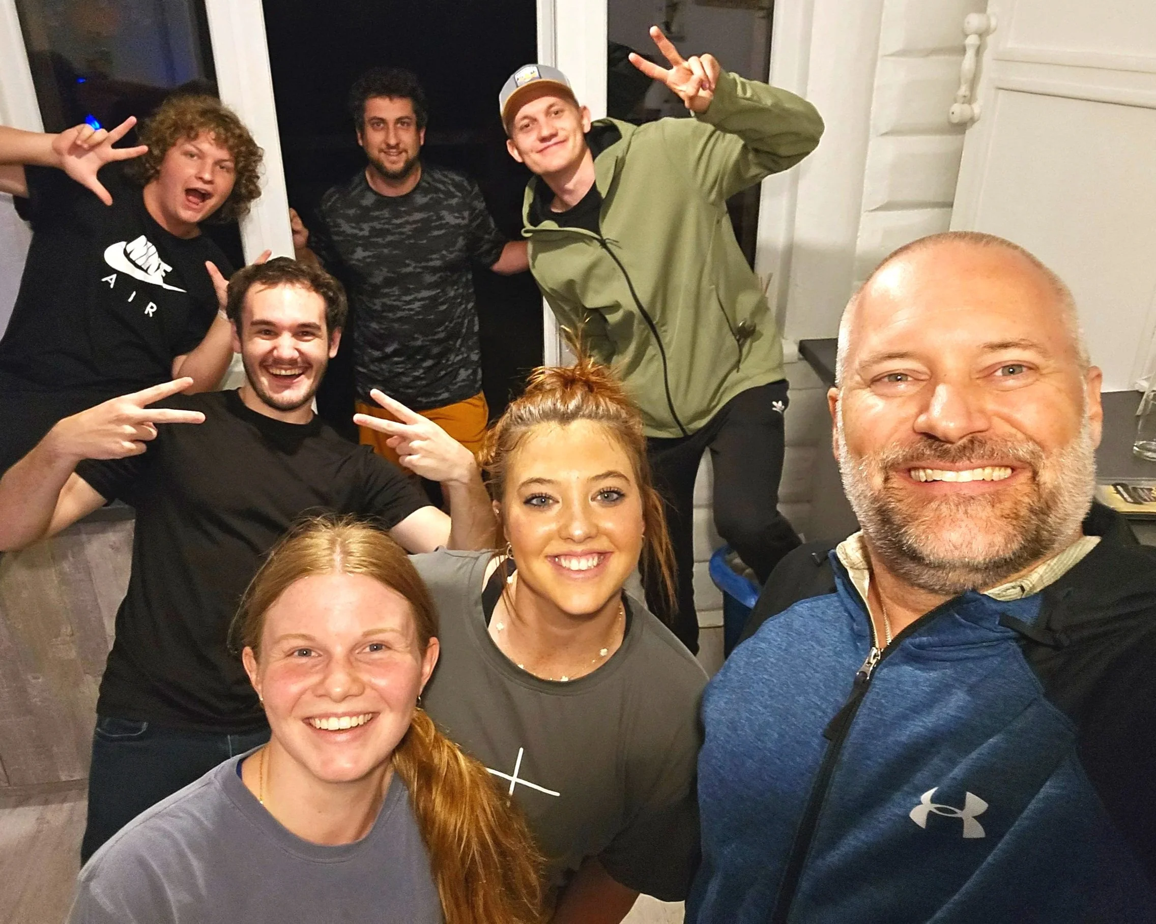 Group of eight people taking a selfie together, smiling and making peace signs, indoors at night. They are ready to start a week of camp overseas.