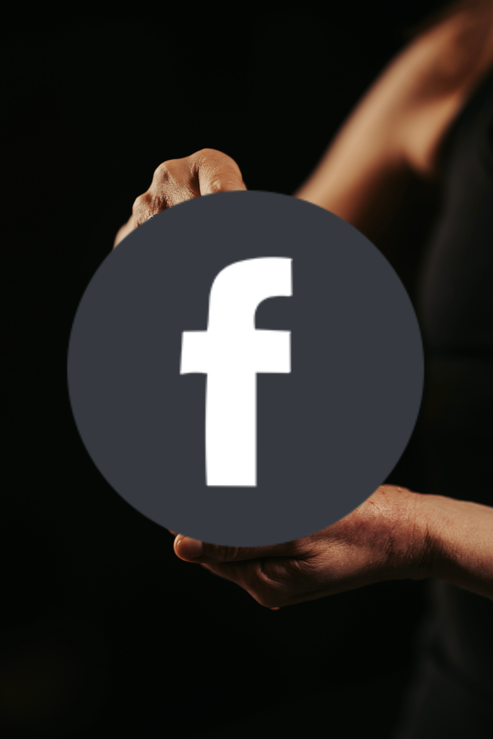 A person holding a circular Facebook logo in front of their body against a dark background.