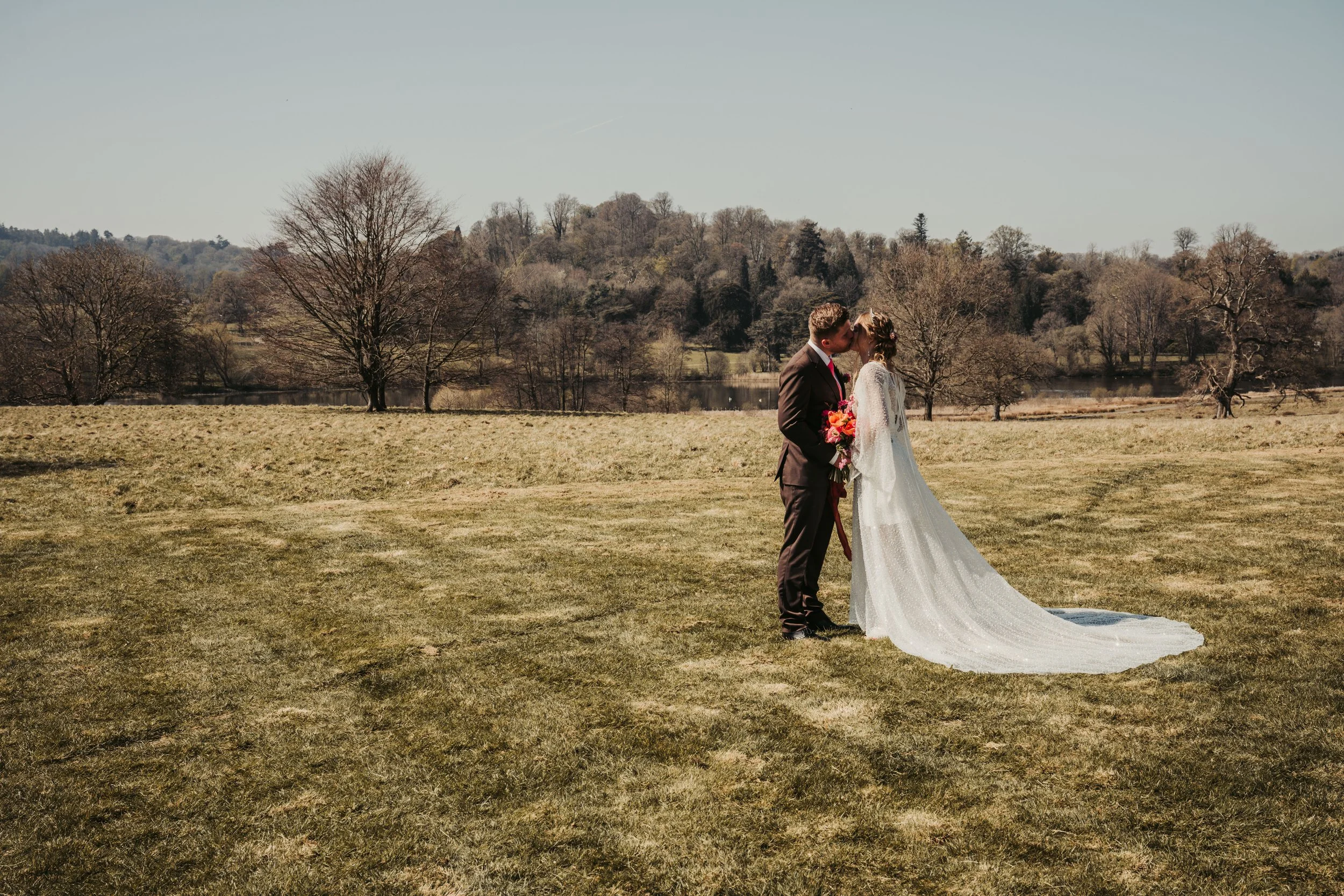 A couple in wedding attire sharing a kiss outdoors in a grassy field with trees and a lake in the background.