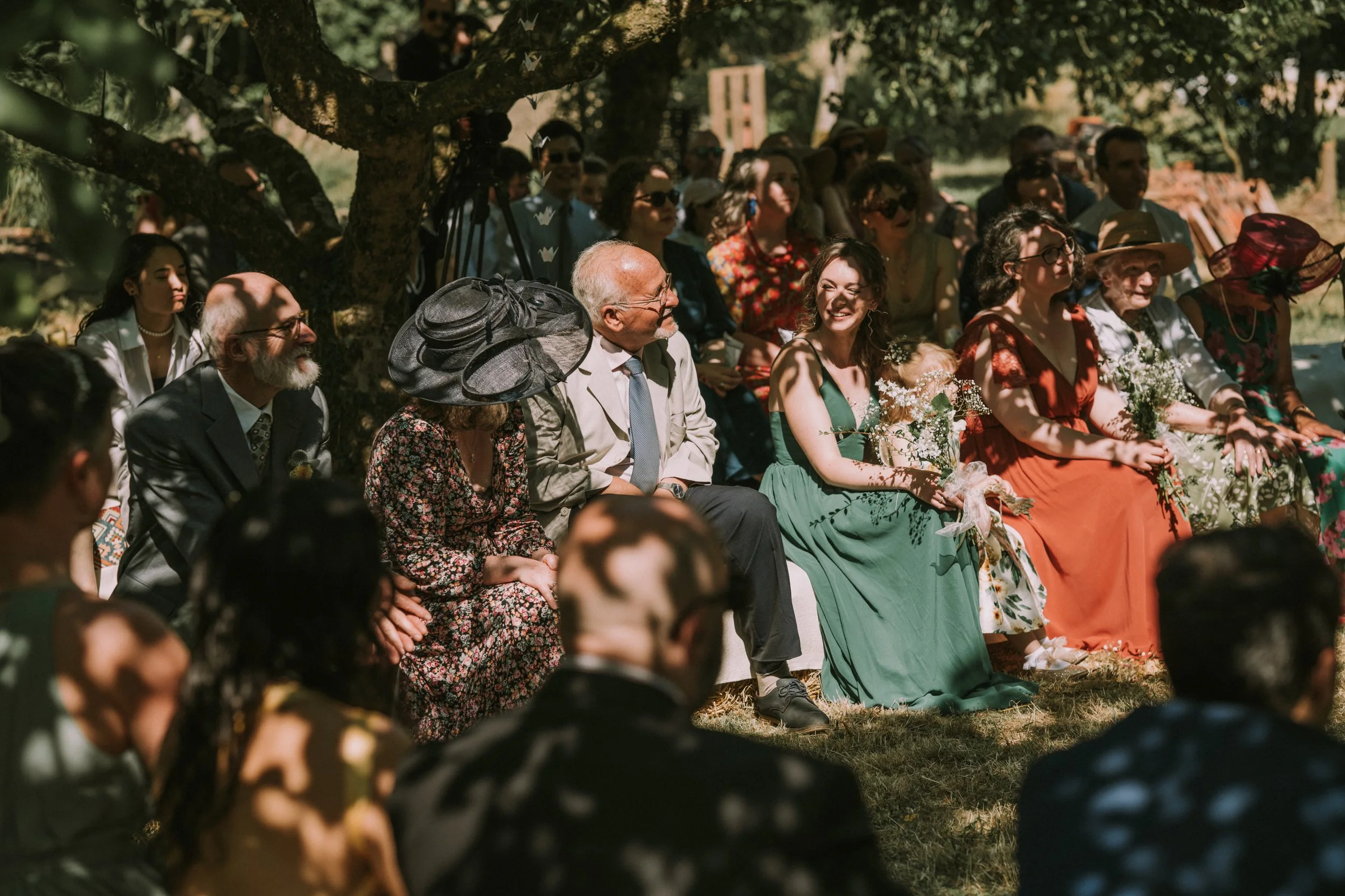 People seated outdoors under a tree during a celebration or wedding, some holding flowers, some wearing hats and colorful dresses.