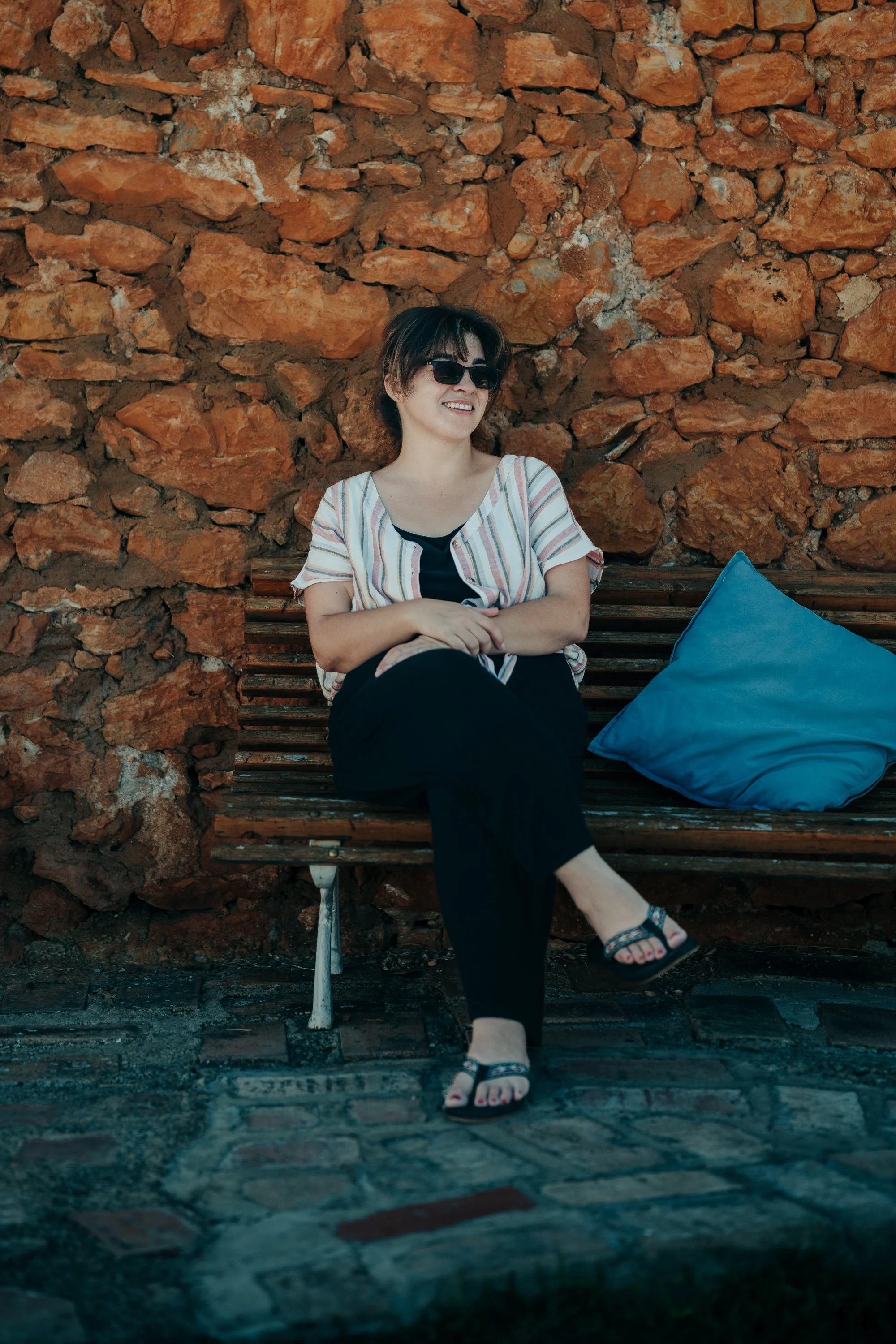 A woman wearing sunglasses, a striped shirt, and black pants sitting on a wooden bench with a blue pillow, against a brick wall background.