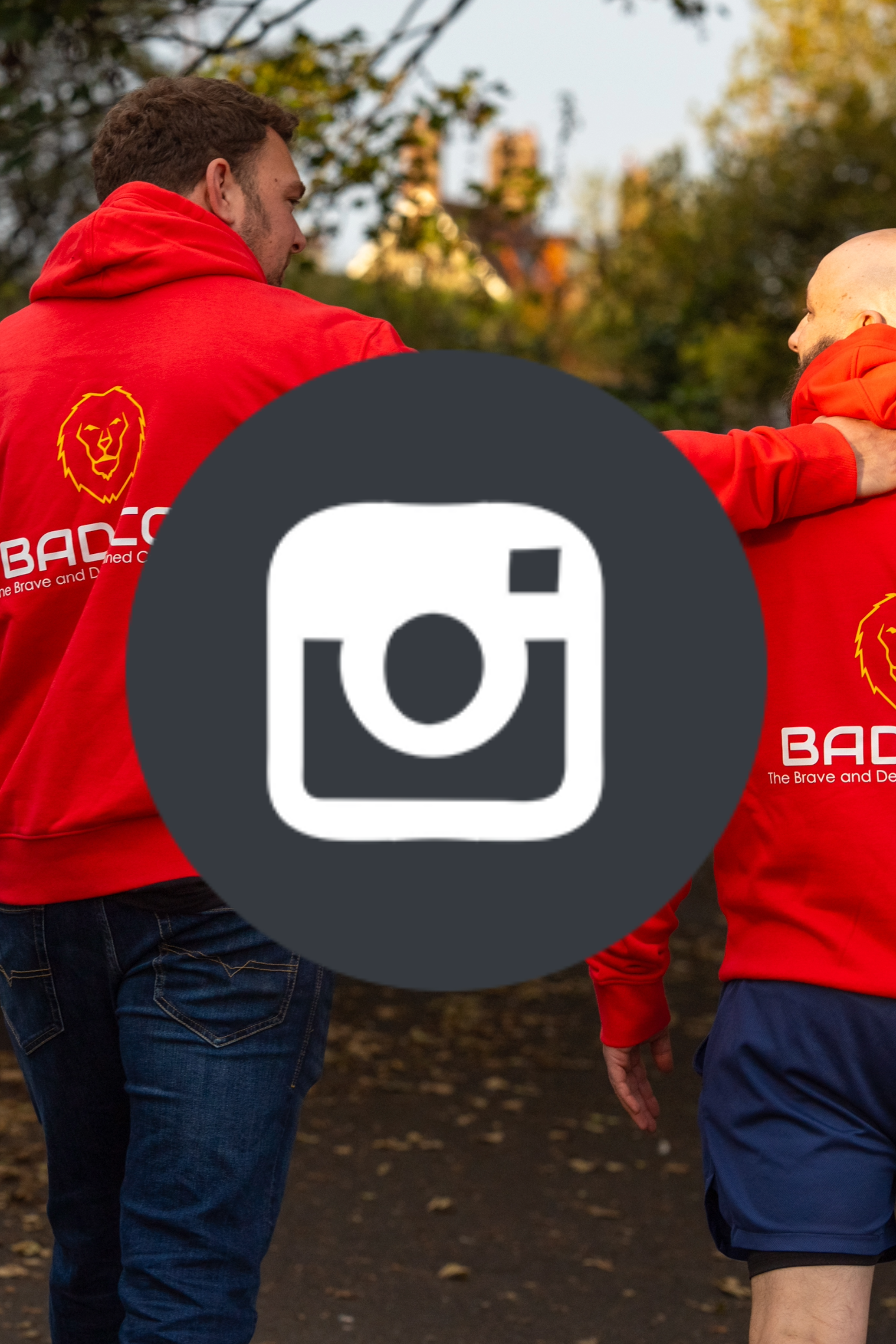 Two men wearing red hoodies with a lion logo and the word 'BAD' on the back, walking together outdoors in a park with autumn trees.