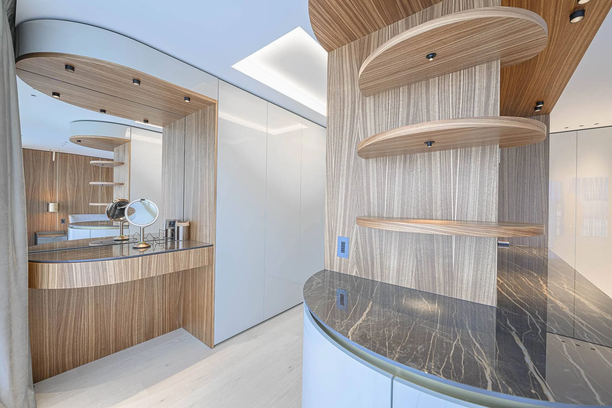 Intérieur moderne avec des murs en bois clair, un comptoir en marbre noir avec des veines dorées, étagères en bois courbes, et un miroir rond sur une coiffeuse.