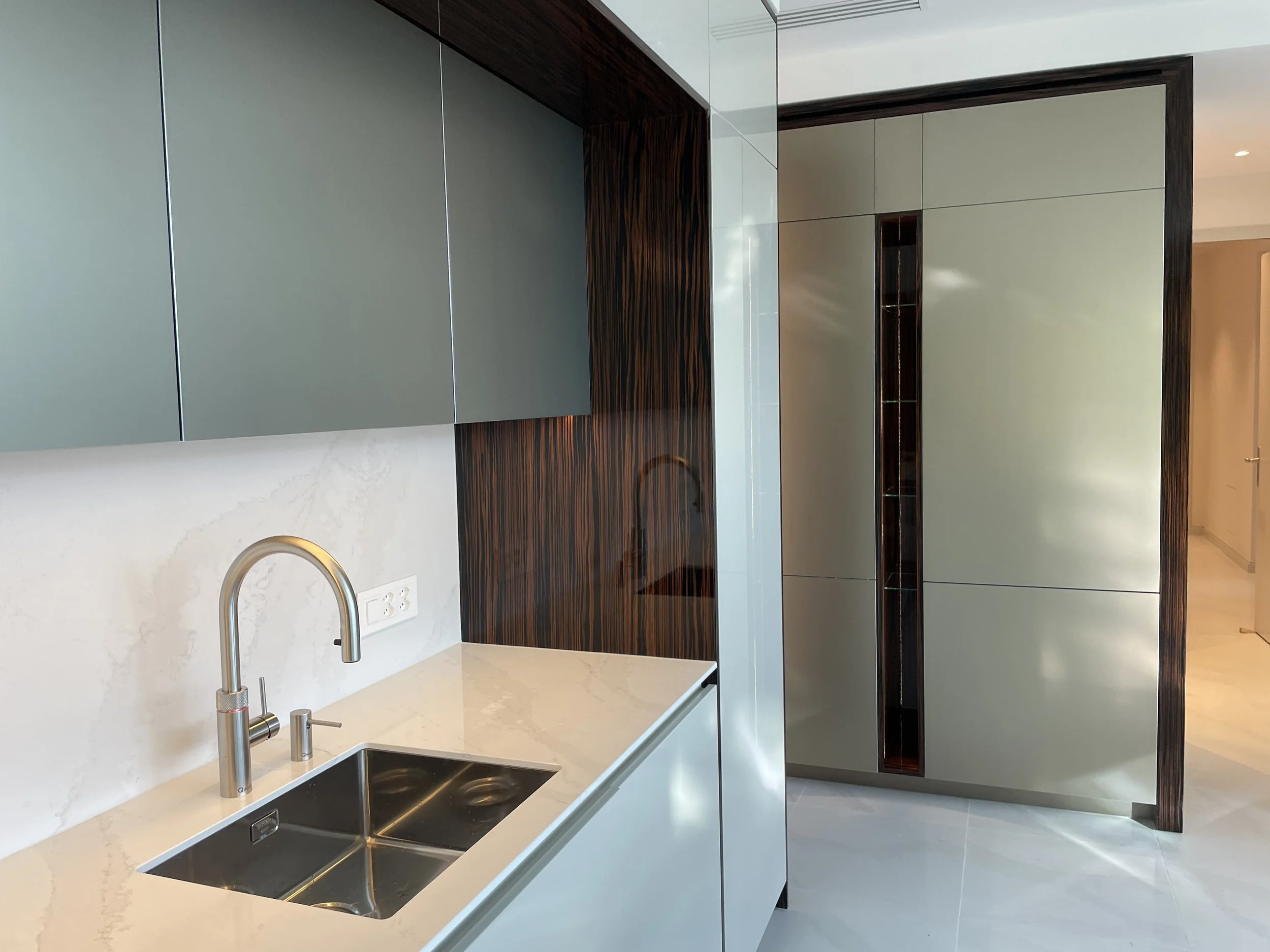 Une cuisine moderne avec un évier en acier inoxydable, un robinet, des armoires en gris et du bois foncé, et un sol en carrelage clair.