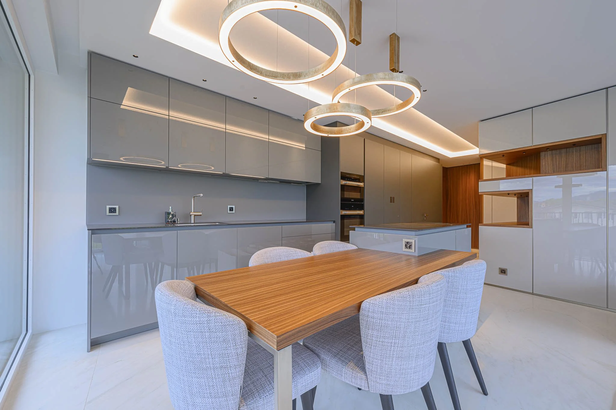 Cuisine moderne avec un plan de travail gris, armoires brillantes, et luminaires suspendus en forme d'anneaux, avec une table en bois et des chaises en tissu clair
