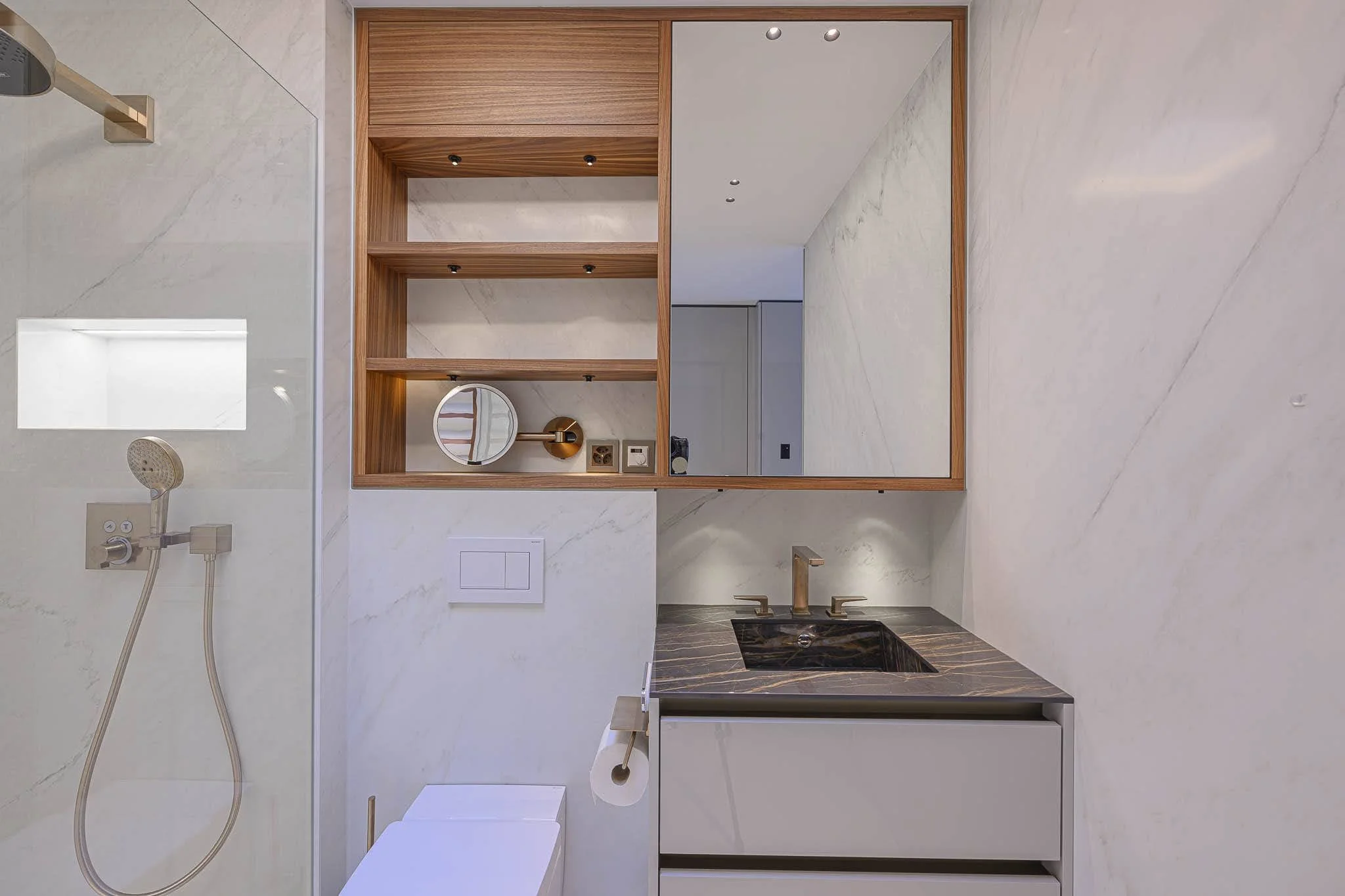 Salle de bain moderne avec une douche à gauche, lavabo avec comptoir en pierre noire, grand miroir, étagères en bois, murs en marbre blanc, toilettes et robinets en laiton