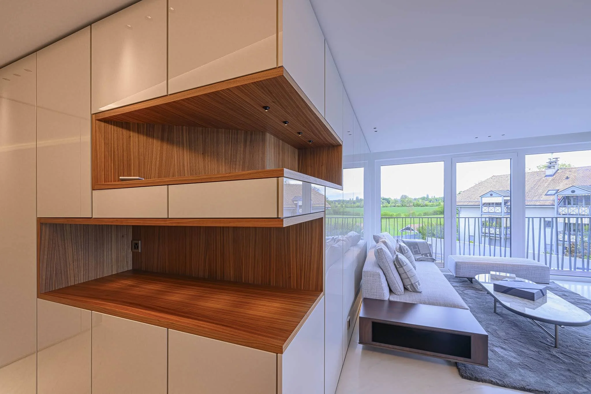 Salon moderne avec canapé beige, fauteuils, table basse en marbre et grandes fenêtres offrant une vue sur un espace vert et un bâtiment adjacent.