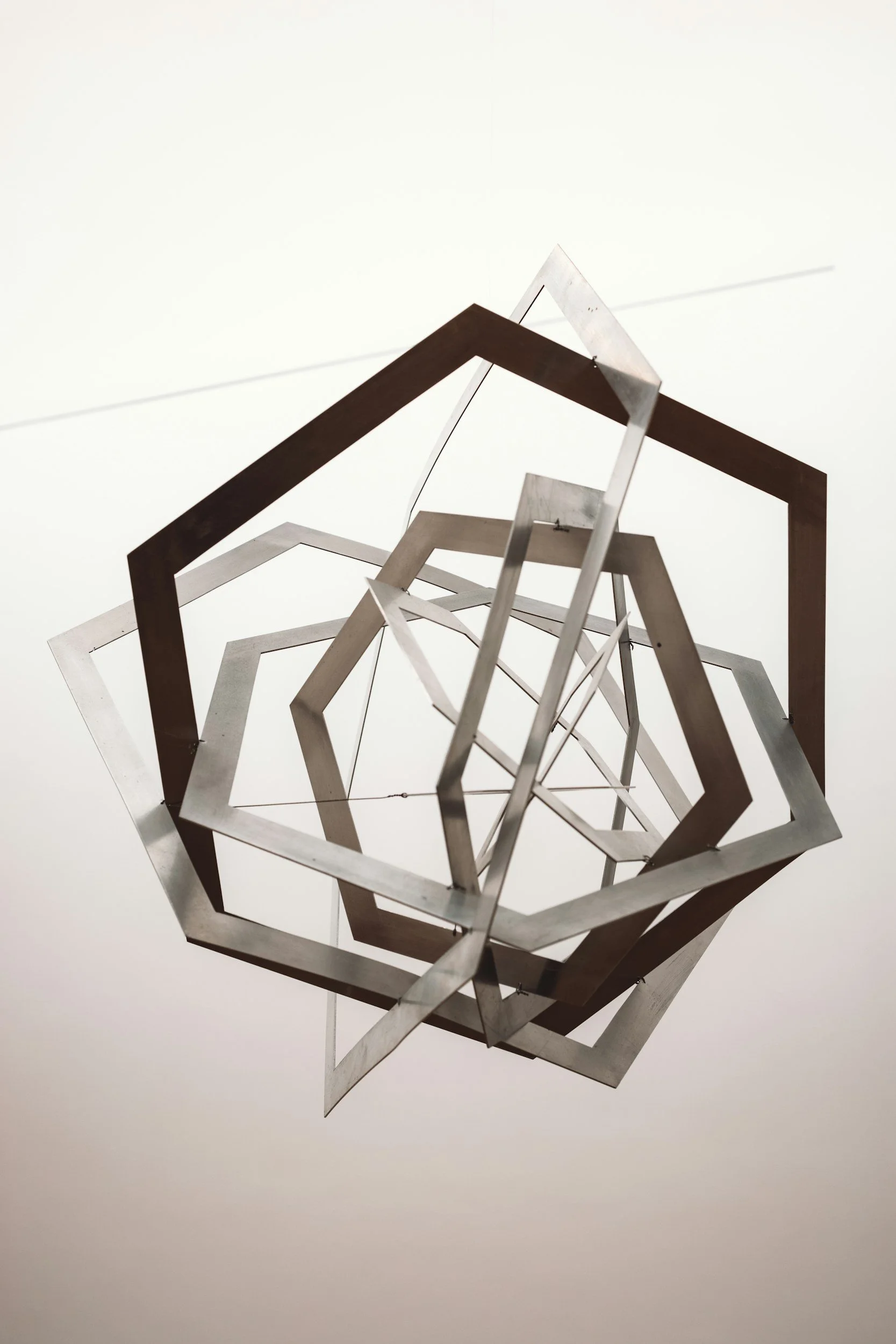 A geometric metal sculpture hanging from the ceiling, consisting of interconnected pentagon-shaped frames in shades of silver and bronze, viewed from below against a plain white ceiling.