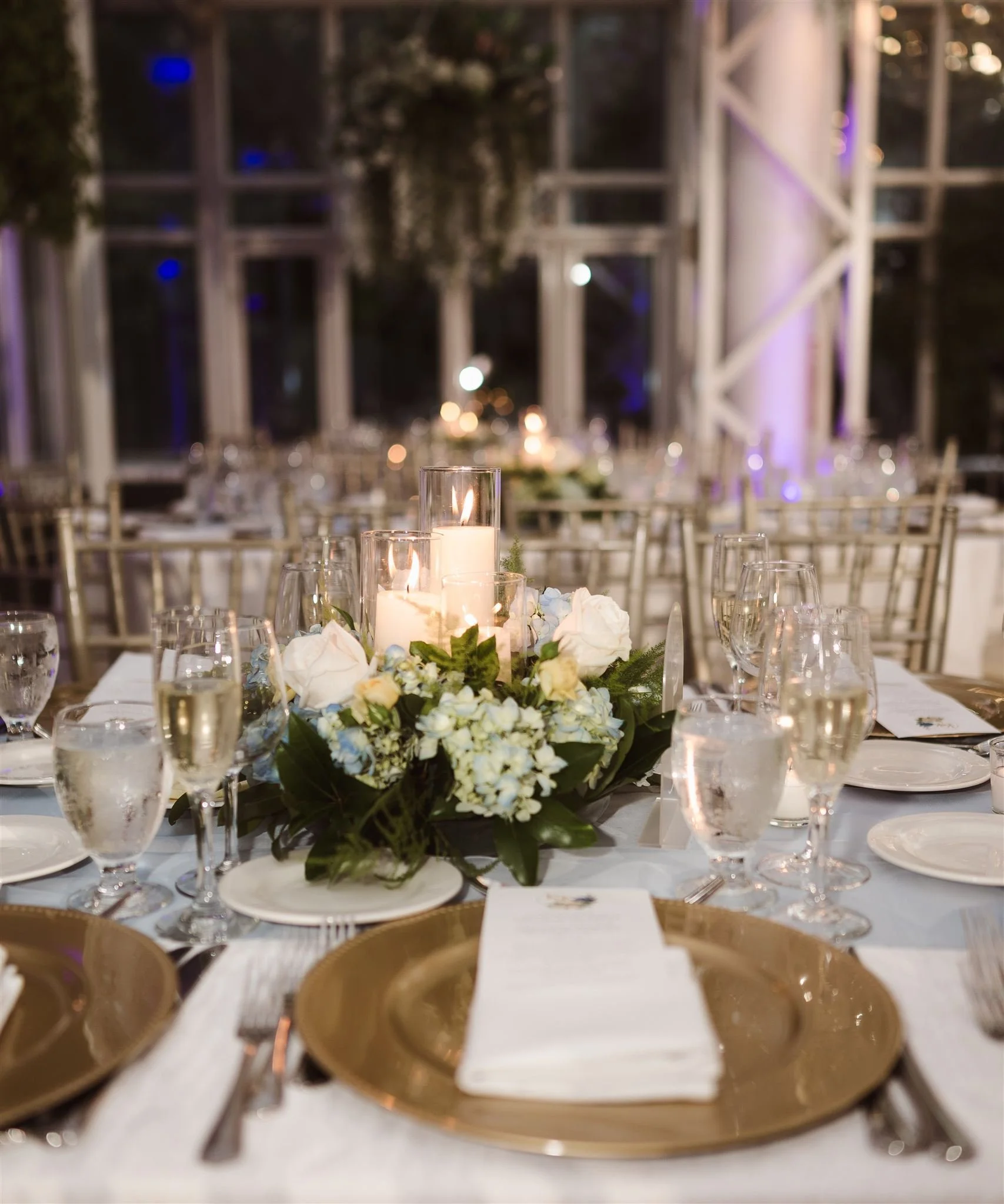 A beautifully decorated banquet table with floral centerpiece, white napkin, gold charger plates, wine glasses, candles, and silverware in an elegant setting.