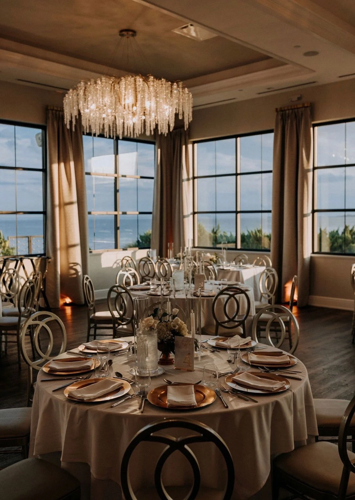 Elegant dining room with round tables set for a formal event, decorated with white floral centerpieces, candles, and gold chargers, large windows showing an ocean view, and a crystal chandelier hanging from the ceiling.