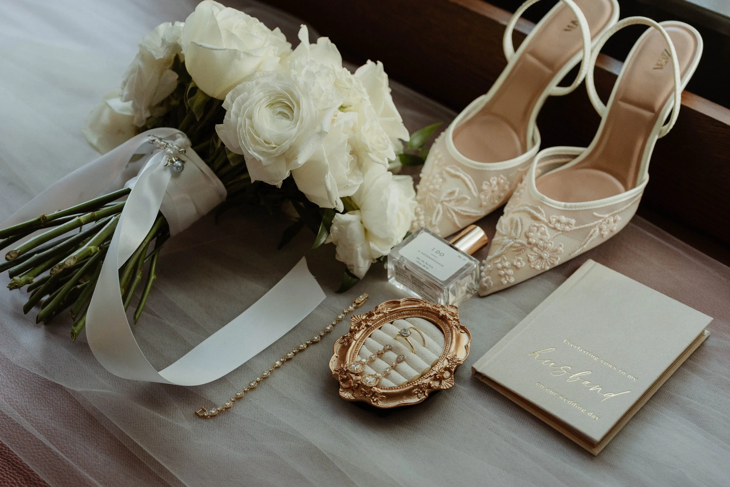 A bridal bouquet of white flowers, a pair of embroidered white bridal shoes, a perfume bottle, a jewelry box with rings, a bracelet, an invitation card, and a small vase of white flowers arranged on a table.