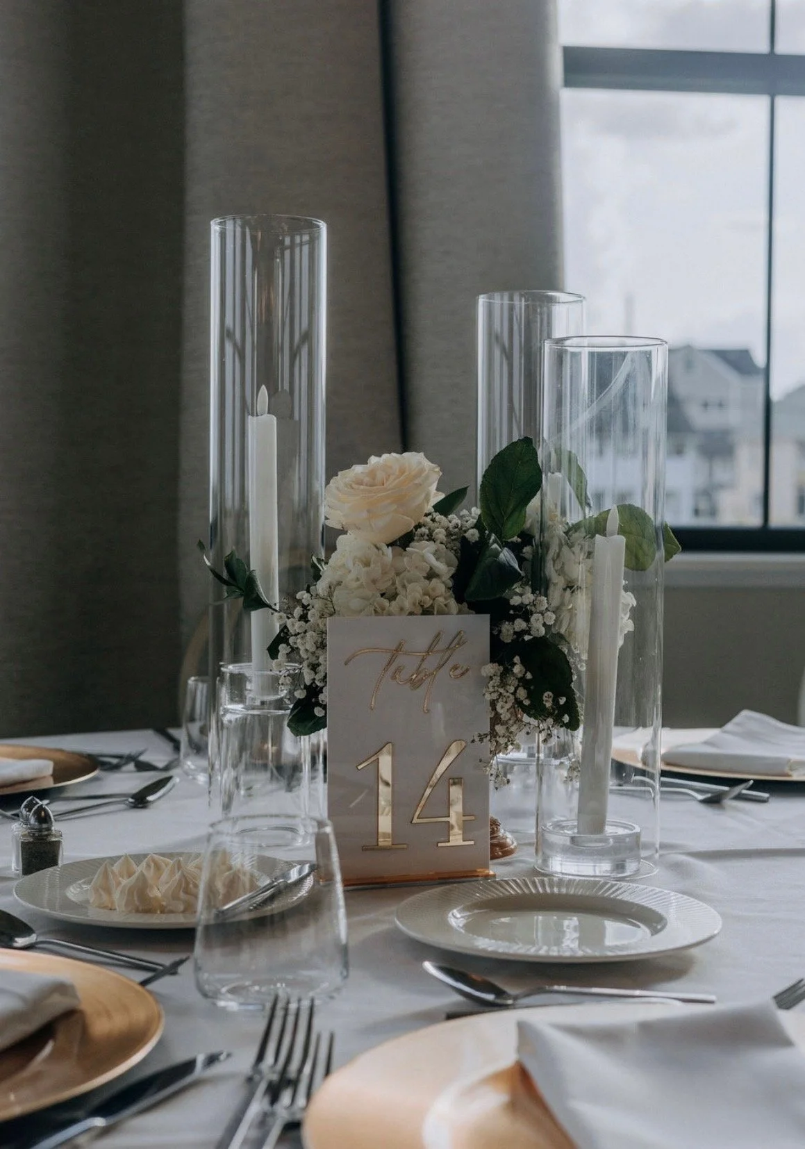 Elegant dining table decorated with white flowers, candles in tall glass holders, and a table card that reads 'Table 14'. The setting includes plates, utensils, glasses, and napkins, with a window in the background.