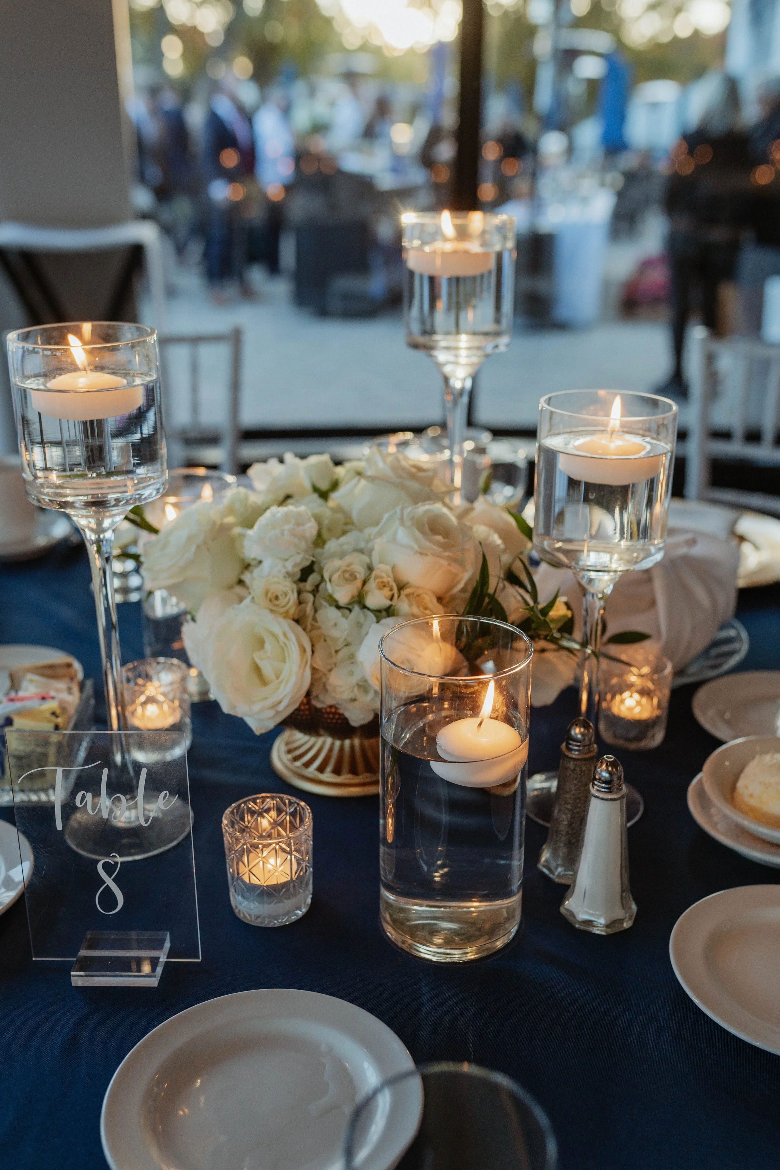 Wedding reception table decorated with white flowers, floating candles in glass holders, and a dark tablecloth. A sign reads 'Table 8'.