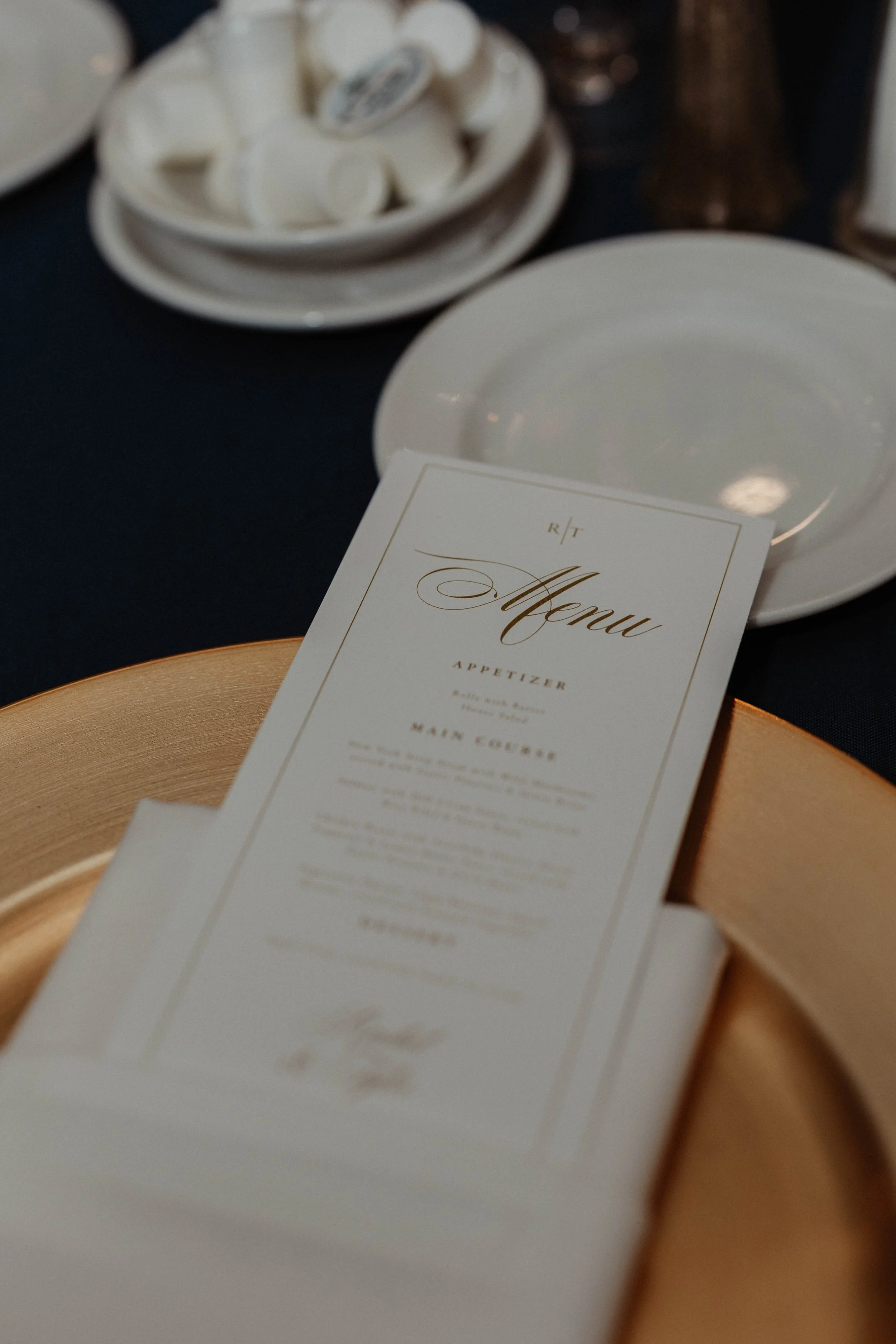 Close-up of a printed menu on a white napkin, placed on a gold-colored charger plate, with a white dish and small white bowls in the background.