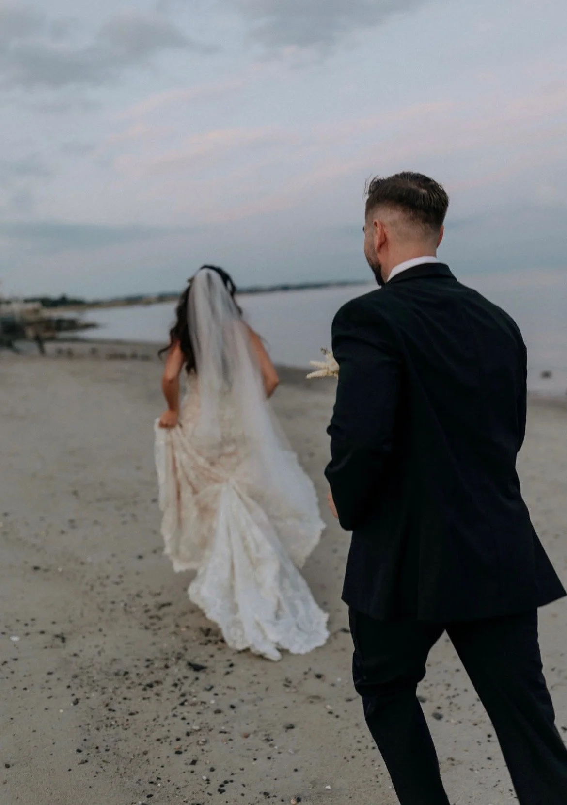 A man in a black suit holding a bouquet stands on a sandy beach, looking at a woman in a white wedding dress with a train and veil, walking or running towards the water, during a cloudy evening.