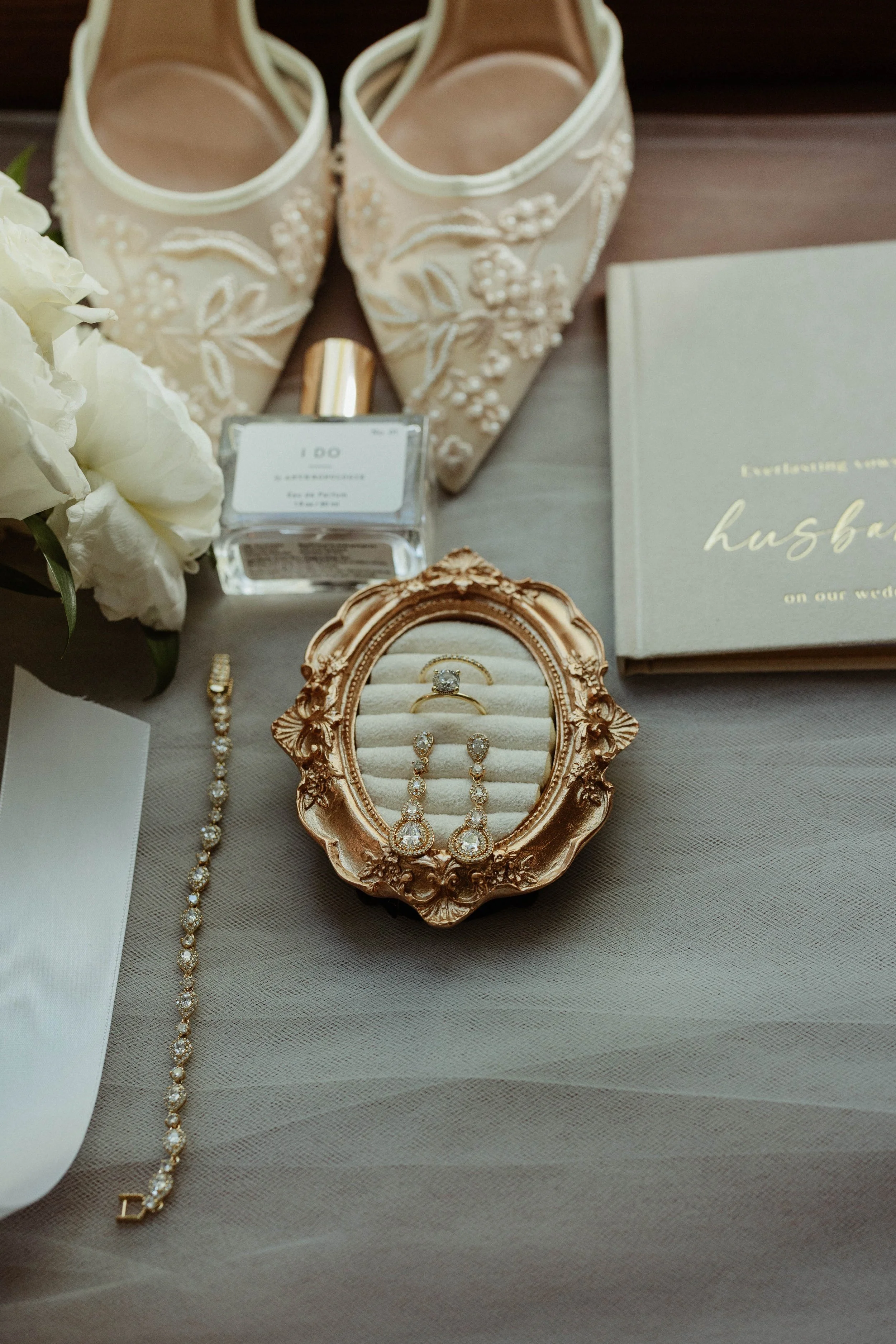 Wedding accessories displayed on a table, including white embroidered bouquets, a bottle of perfume, a book titled 'Husband' in gold lettering, a pair of white lace high-heel shoes, a gold jewelry box with diamond earrings and a ring, and a matching diamond bracelet.