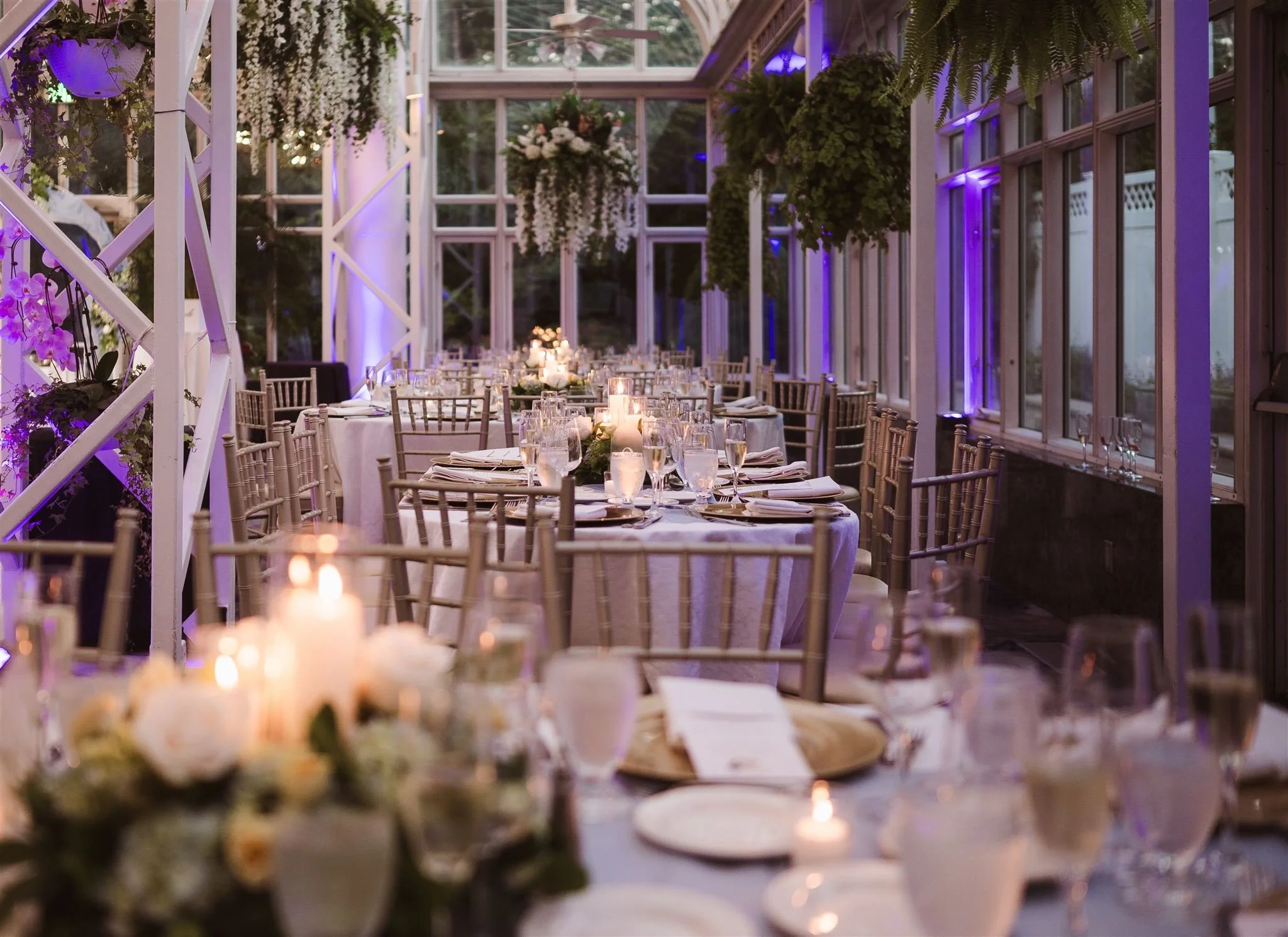 Elegant event space with round tables, decorated with candles, floral arrangements, and glassware, surrounded by wooden chairs with a backdrop of large windows, greenery, and purple lighting accents.